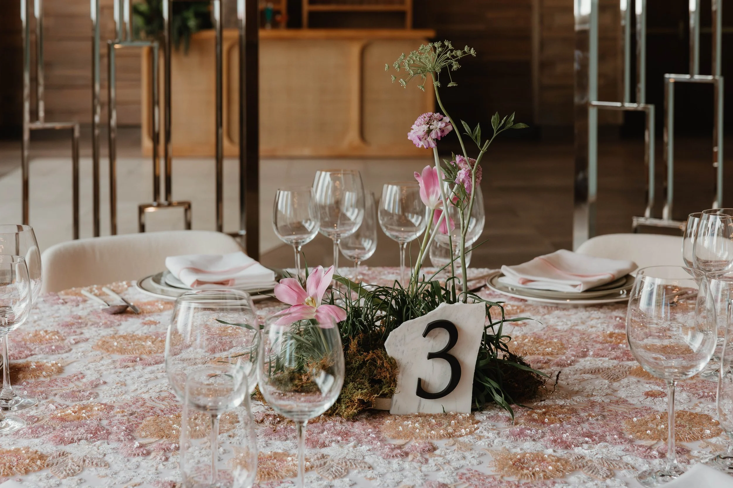 Elegant table setting with pink floral centerpiece, wine glasses, plates with napkins, and a table number 3 card, in a warmly lit room with wooden accents.