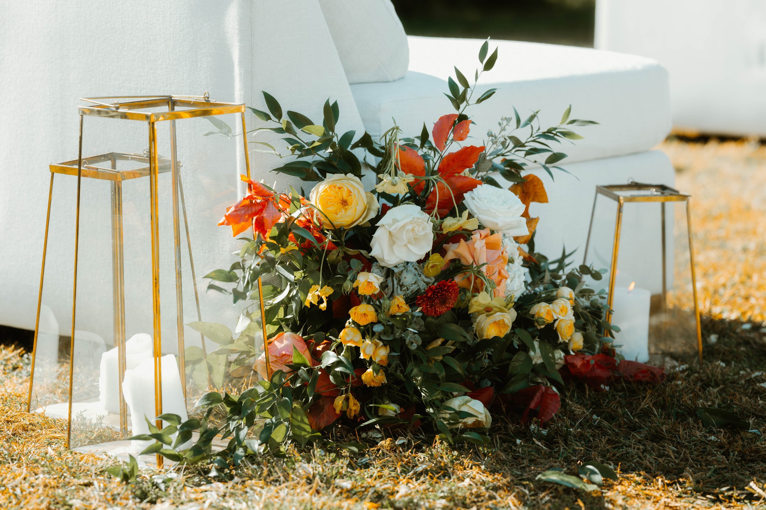 Arrangement of flowers including roses and leaves on the ground beside large glass candle holders with candles inside, against a white sofa. At Chateau Elan