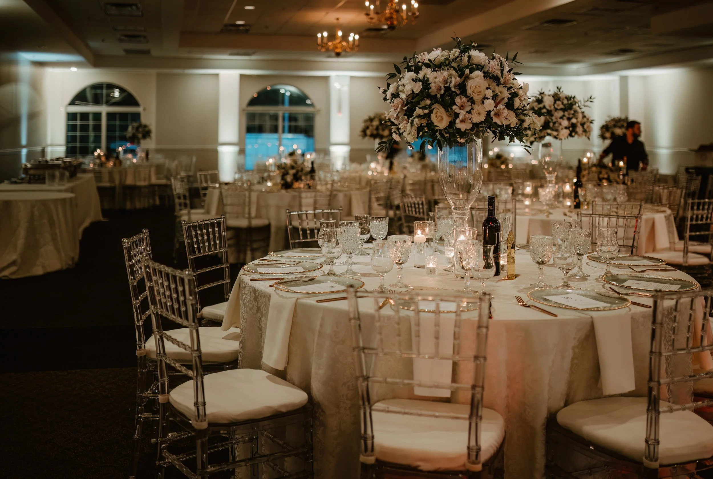 Wedding reception hall with round tables decorated with white tablecloths, elegant glassware, and tall floral centerpieces, dim lighting, and chandeliers overhead.