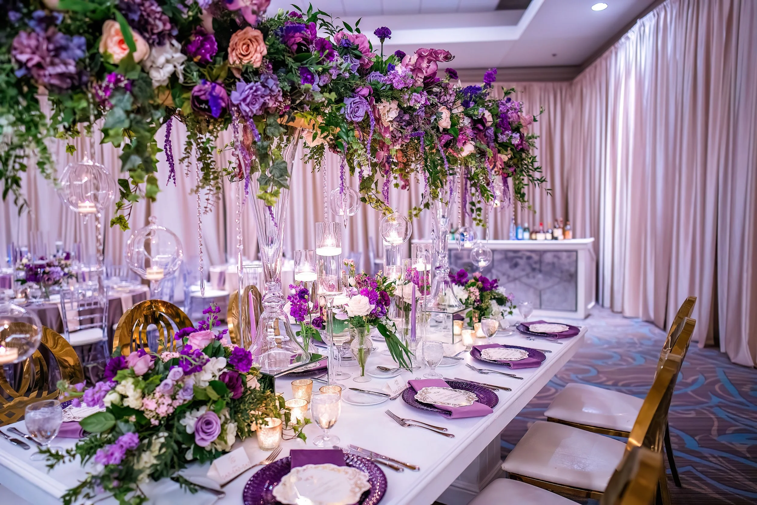 A decorated banquet table with purple and white floral arrangements, tall glass candle holders, and purple napkins, set in an elegant event hall with cream-colored curtains.