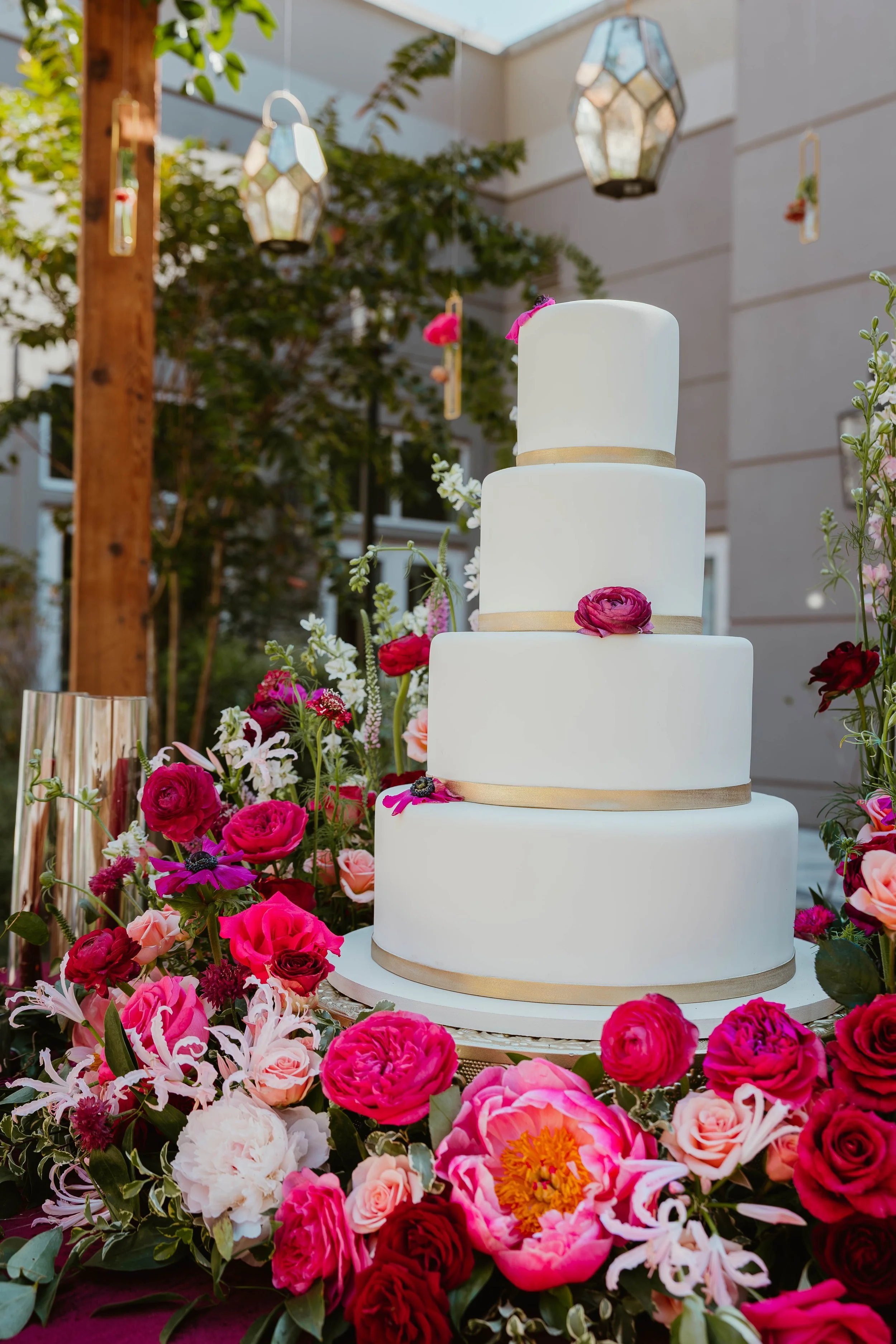 A four-tier white wedding cake decorated with pink, red, and purple flowers, surrounded by a lush floral arrangement, set outdoors with hanging lanterns.