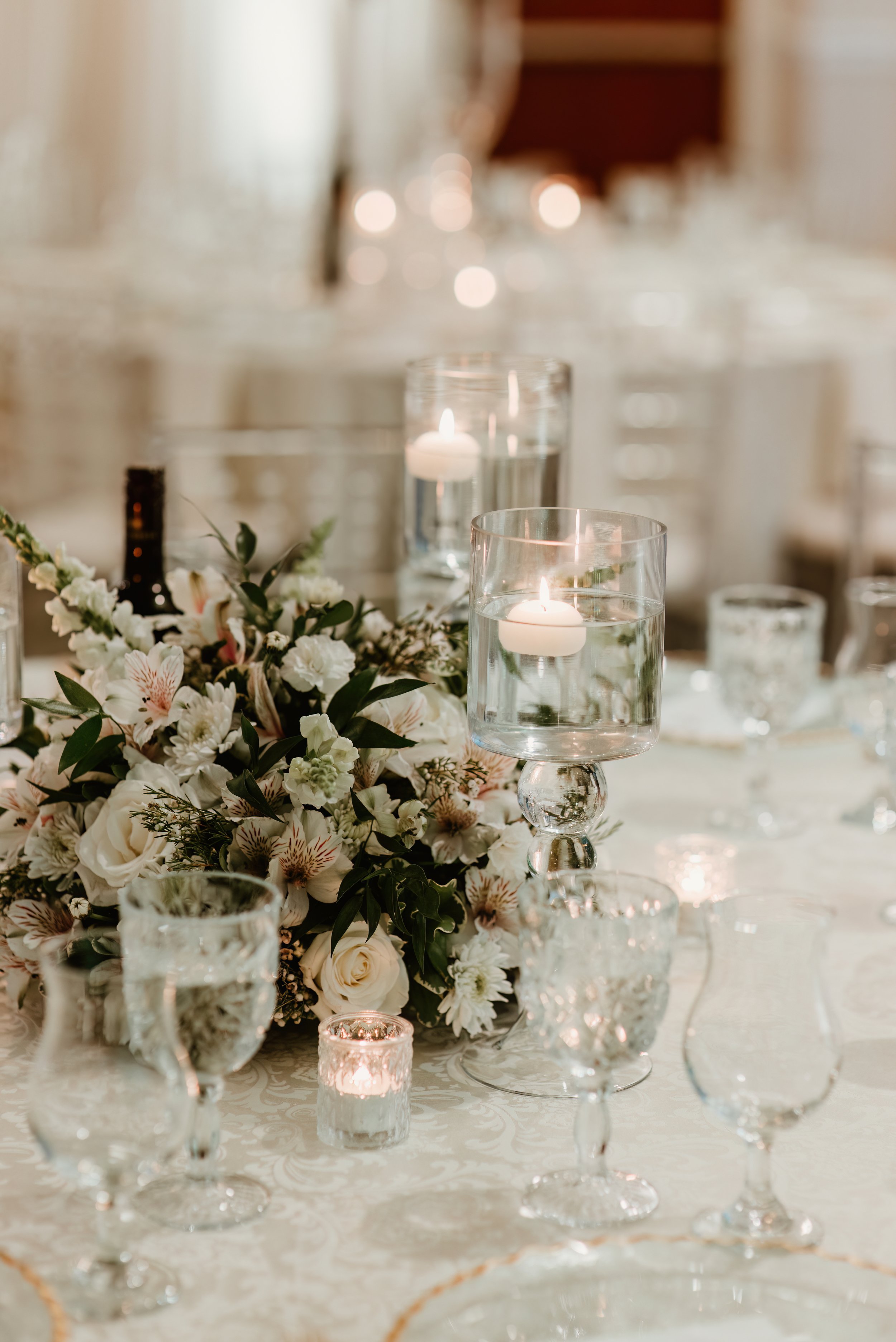 Elegant table centerpiece with white flowers, floating candles in glass holders, and various glassware at a formal event.