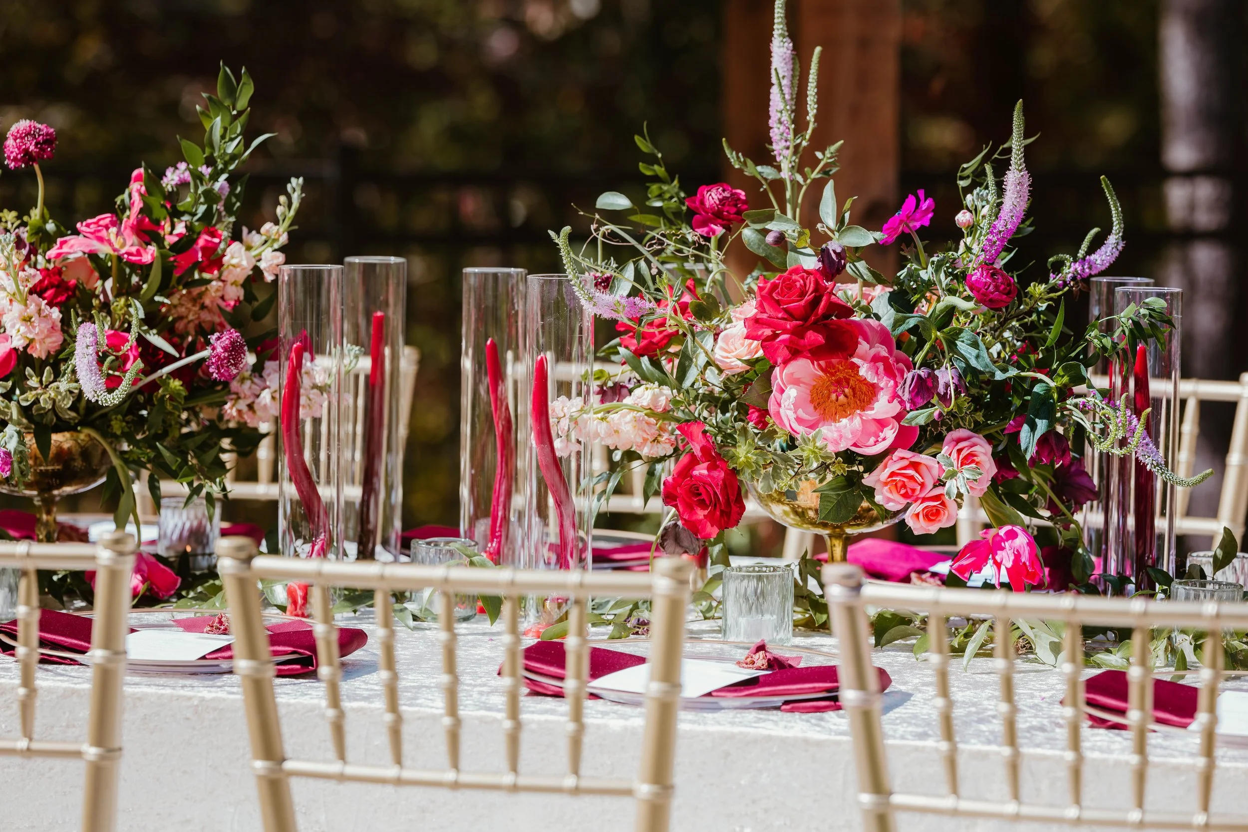 Elegant outdoor dining table decorated with vibrant pink and red flowers in gold vases, tall glass candle holders with red candles, green foliage, and purple accents, set for a special occasion.
