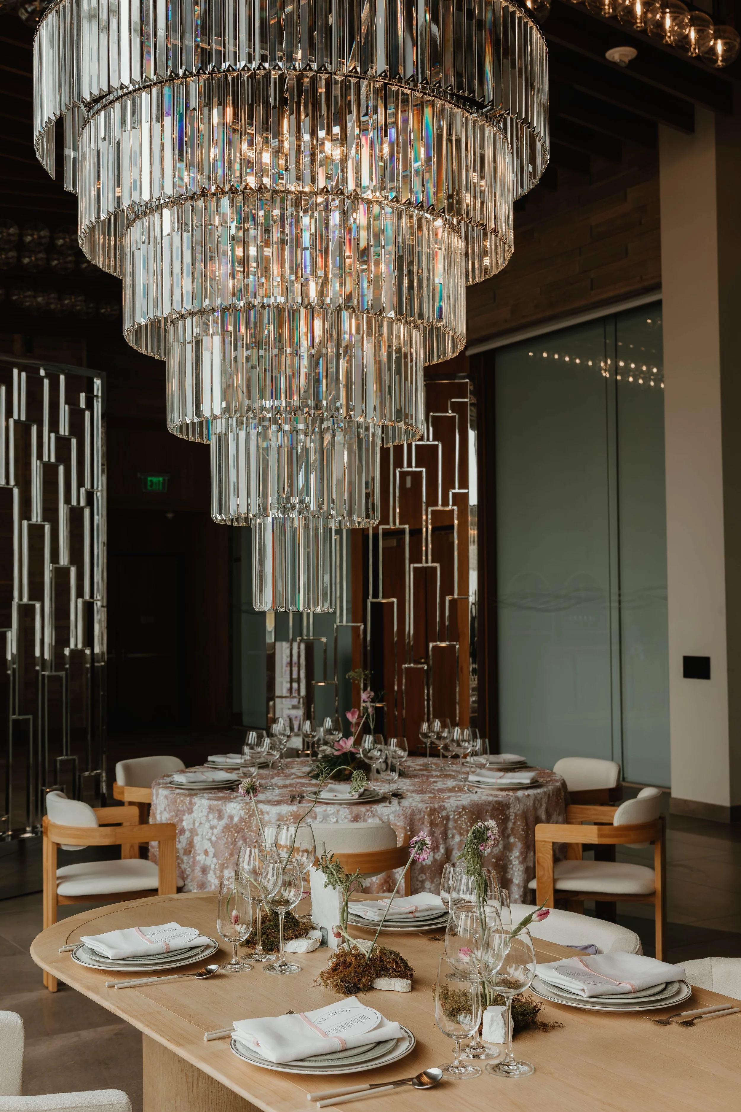 Elegant dining room with large crystal chandelier, round table with floral centerpiece, and neatly set dining arrangements.