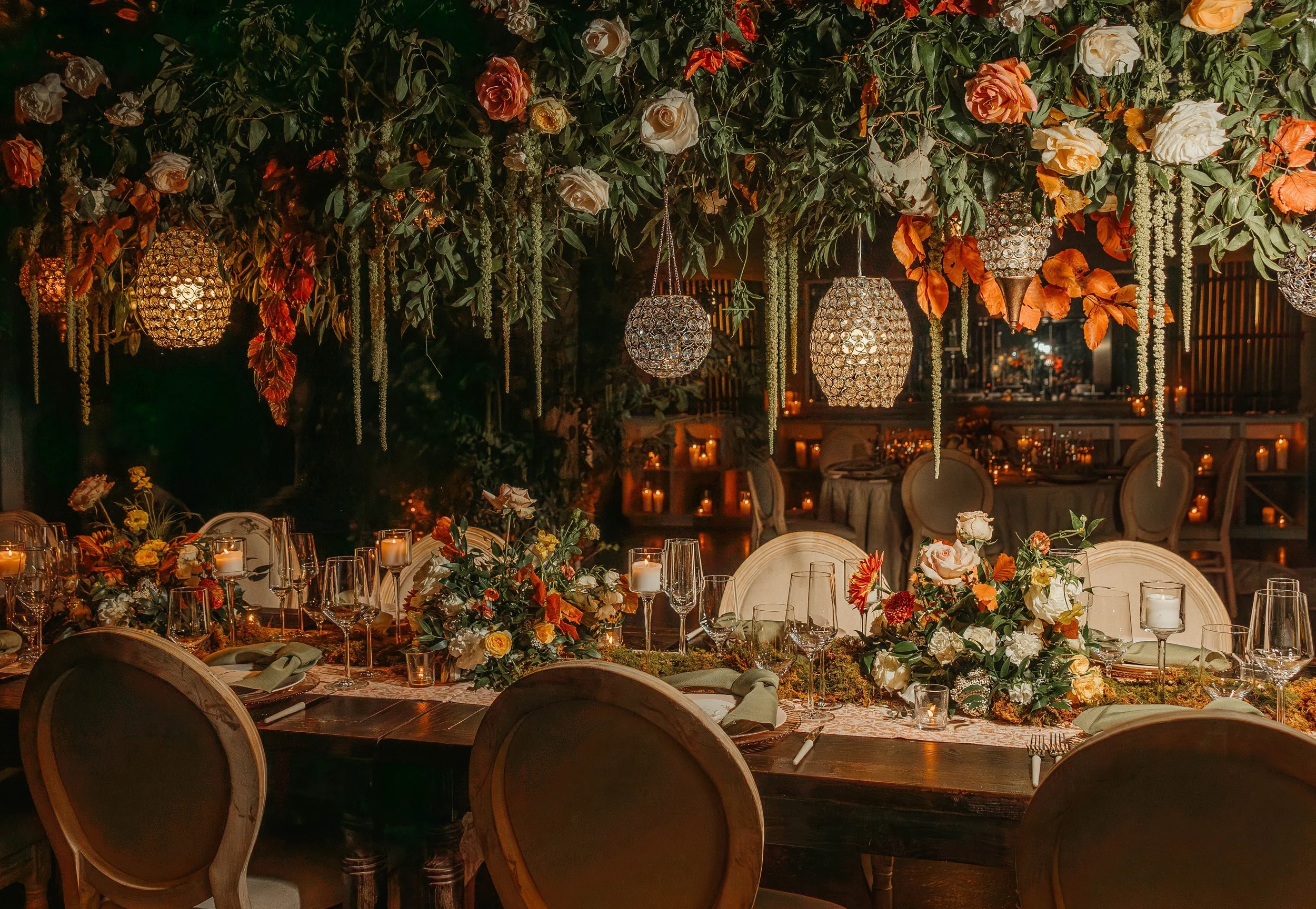 A beautifully decorated banquet table with floral arrangements, candles, wine glasses, and elegant chairs beneath a lush canopy of hanging flowers, greenery, and ornate lamps, set in a warm, intimate setting.