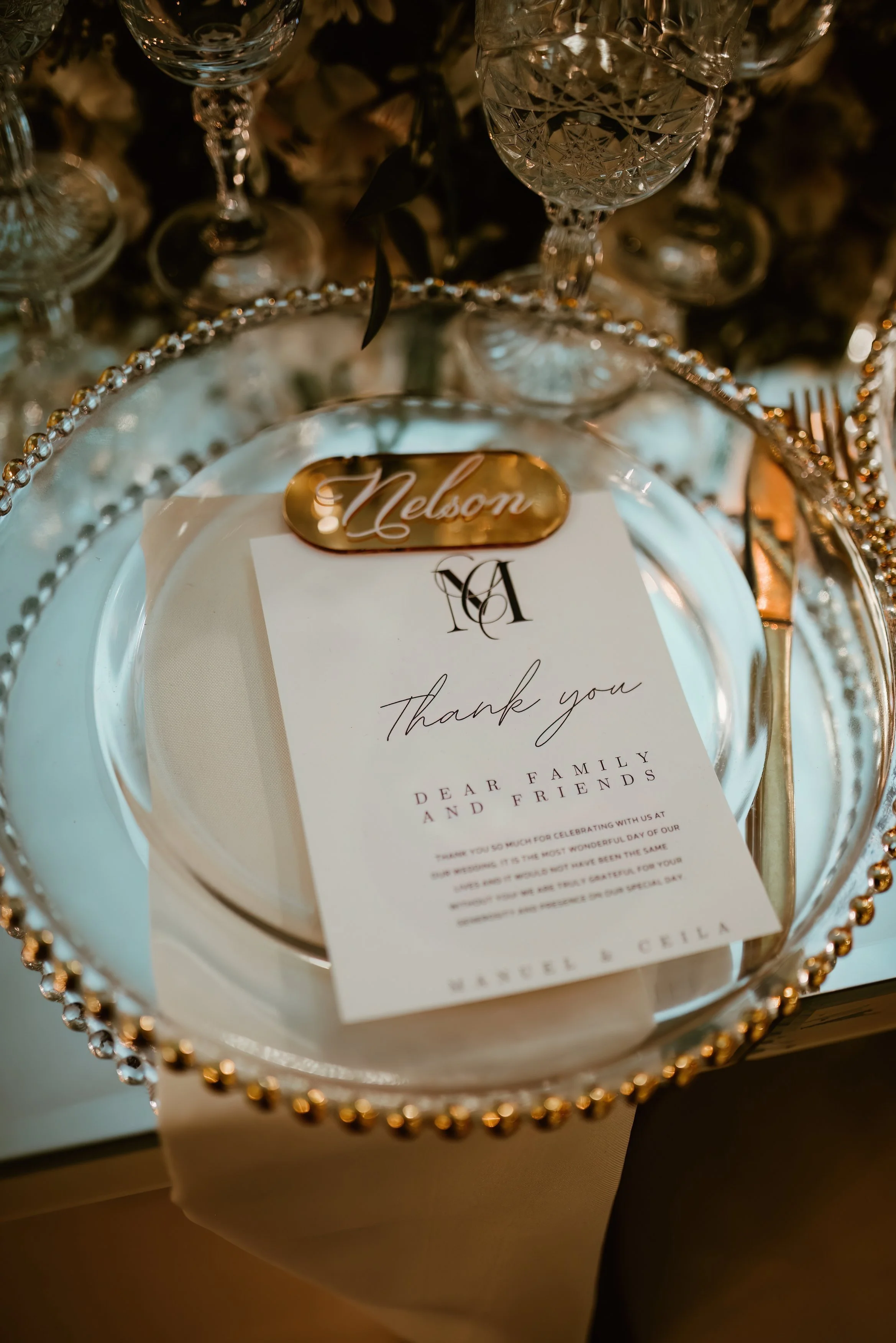 A thank you note placed on a white plate, surrounded by a glass charger and decorative beads, with wedding glasses and flowers nearby.
