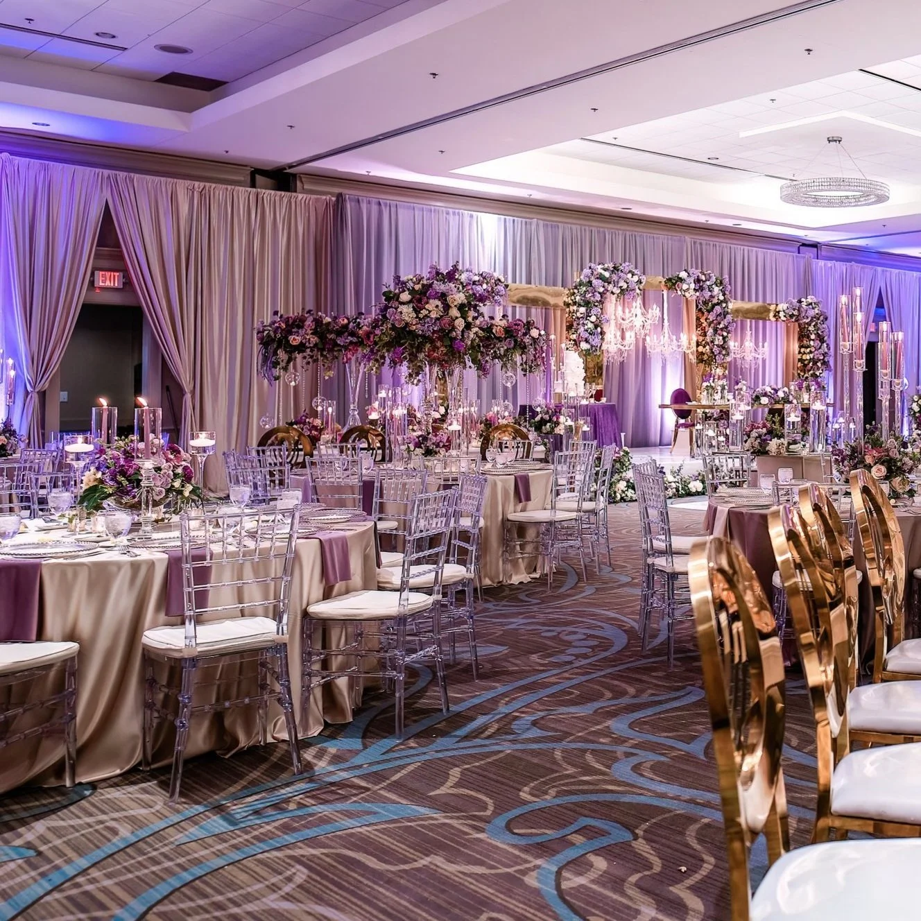Elegant wedding reception setup with floral centerpieces, draped curtains, and a decorated stage with chandeliers in a large banquet hall.