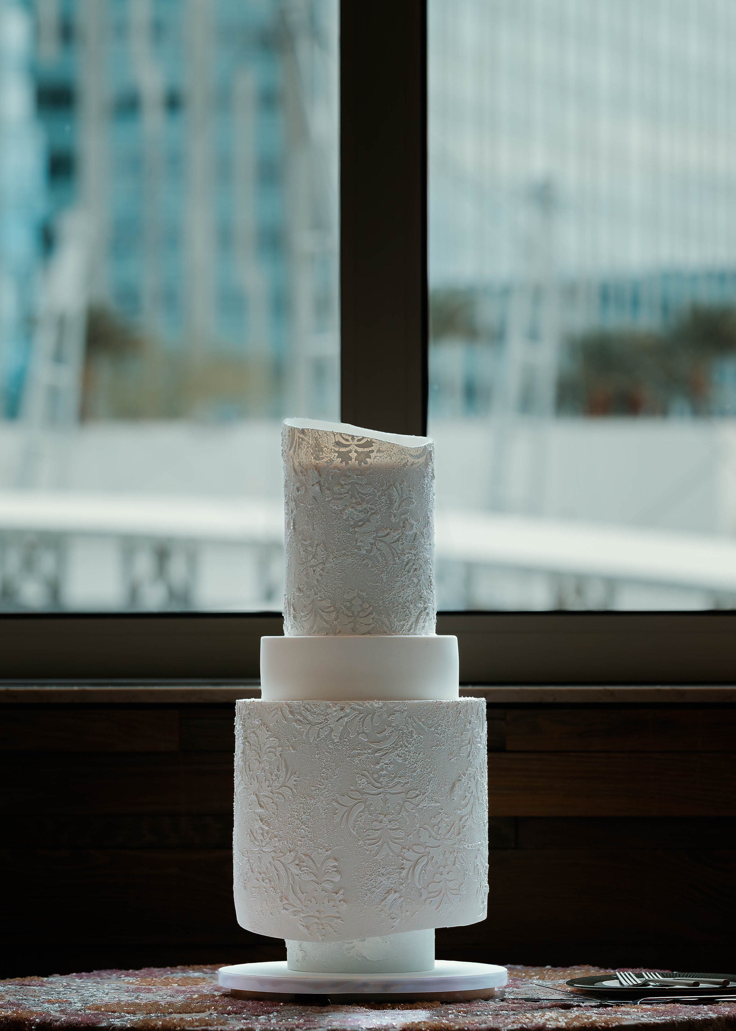 A three-tiered white wedding cake with intricate embossed patterns, placed on a table in front of a window with an urban cityscape background.