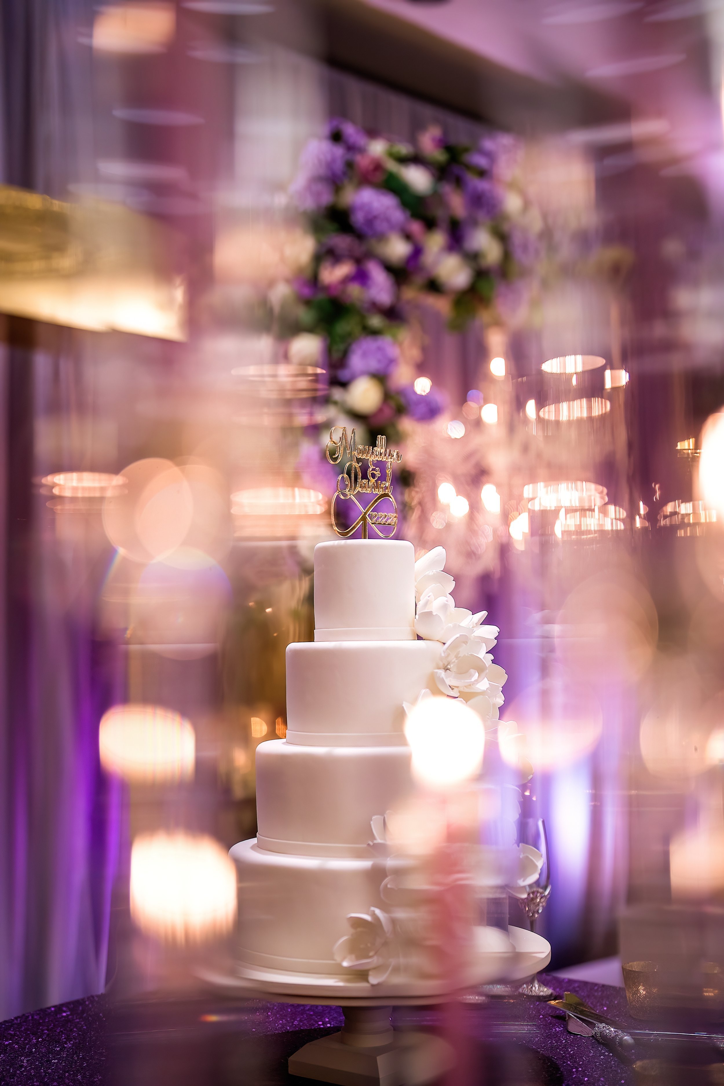 Elegant four-tiered white wedding cake with white floral decorations, topped with a "Happily Ever After" cake topper, against a backdrop of purple and white flowers and warm lighting at a wedding reception.