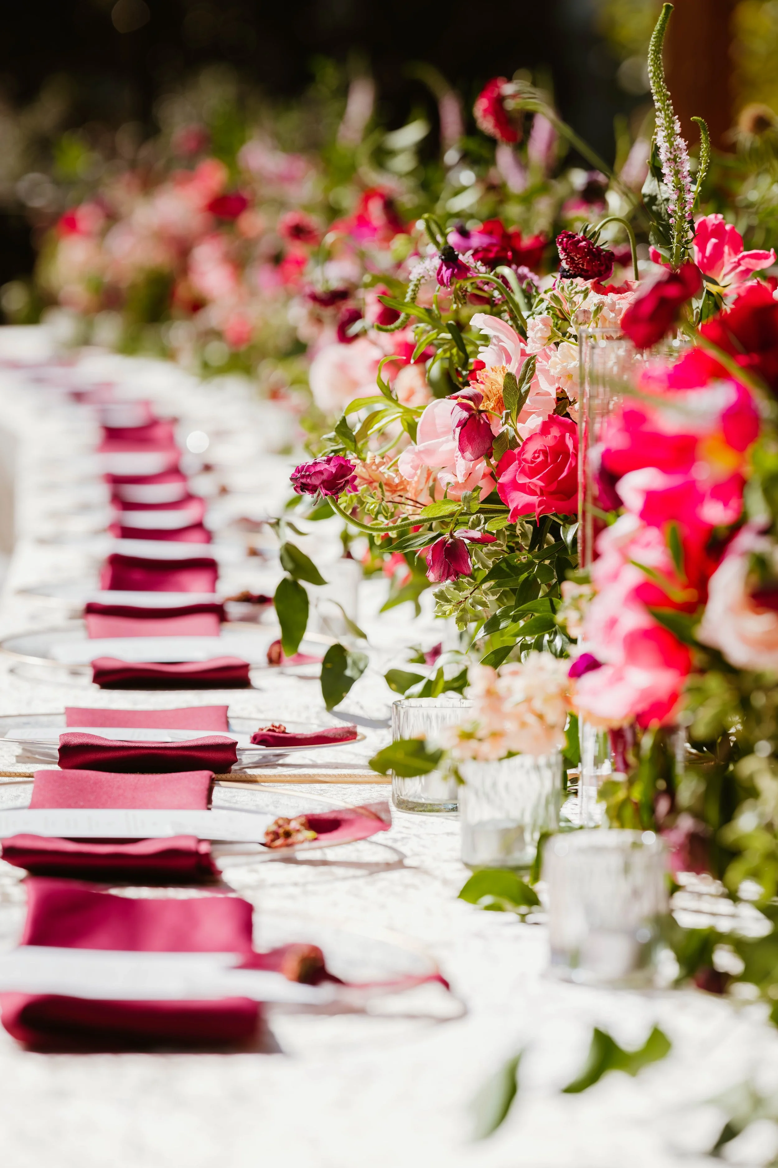 A long outdoor dining table decorated with pink and red flowers and green foliage, set with plates, glasses, and maroon cloth napkins, with sunlight filtering through trees in the background.