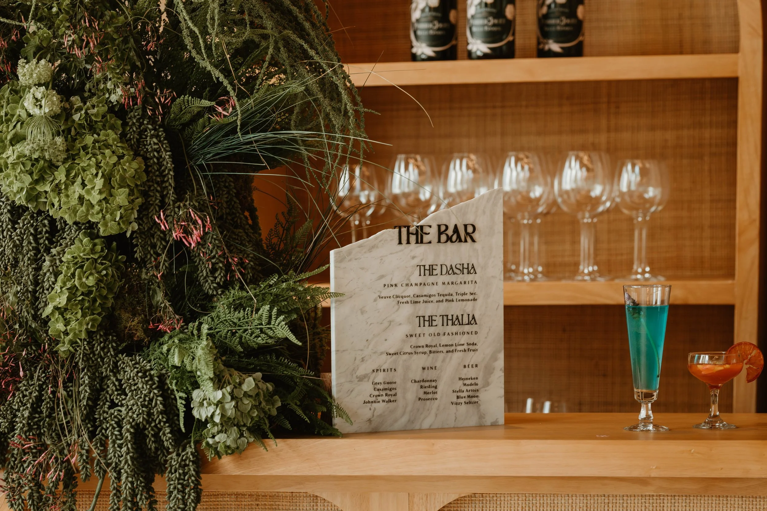 A bar counter with a white marble menu stand and two colorful cocktails, one blue and one orange, in glasses. Behind the counter are empty glasses on a wooden shelf and a green plant arrangement on the left side.