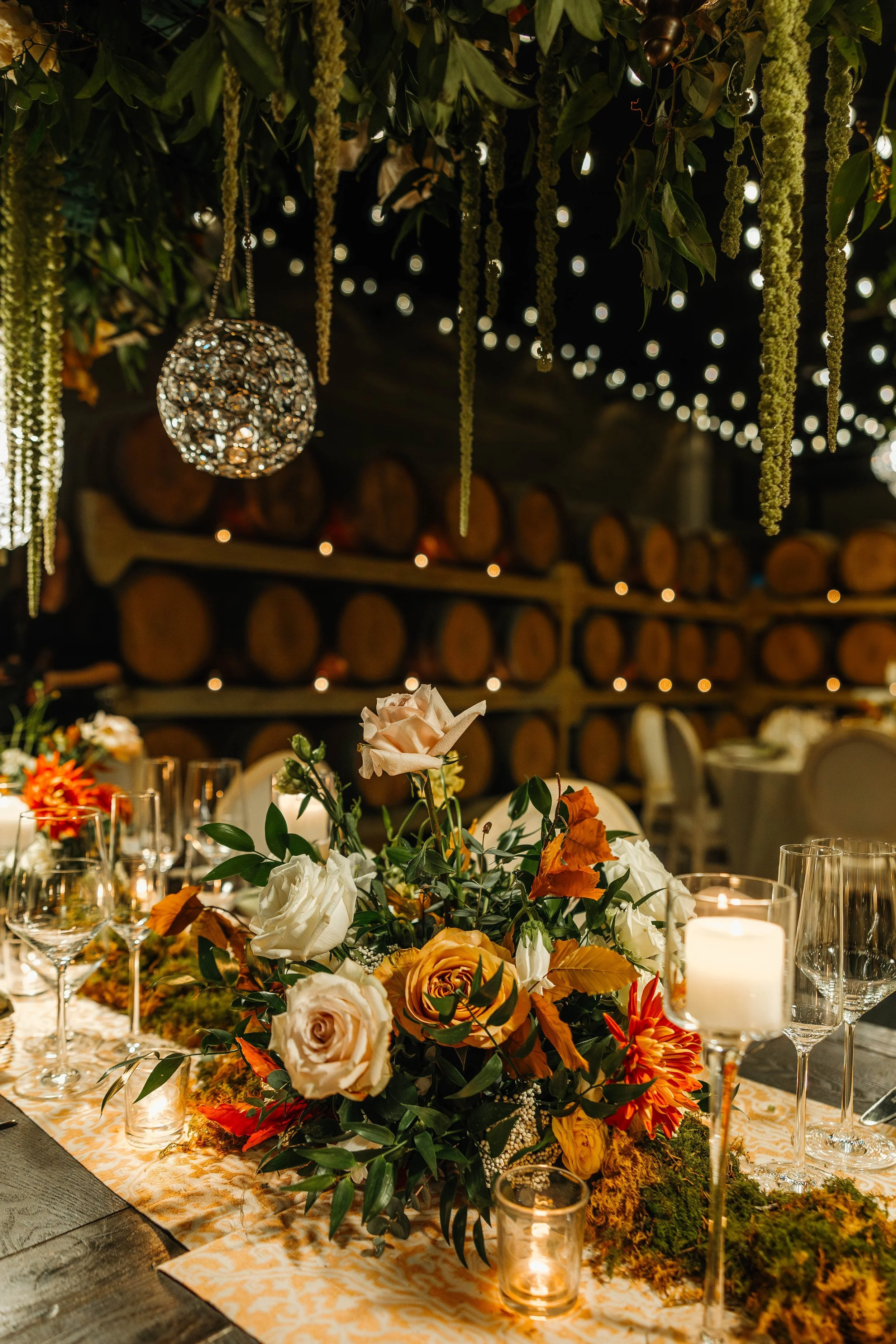 Elegant table decor with a floral centerpiece featuring roses and other flowers, surrounded by candles and wine glasses, with a rustic background of stacked logs and hanging greenery and lights.