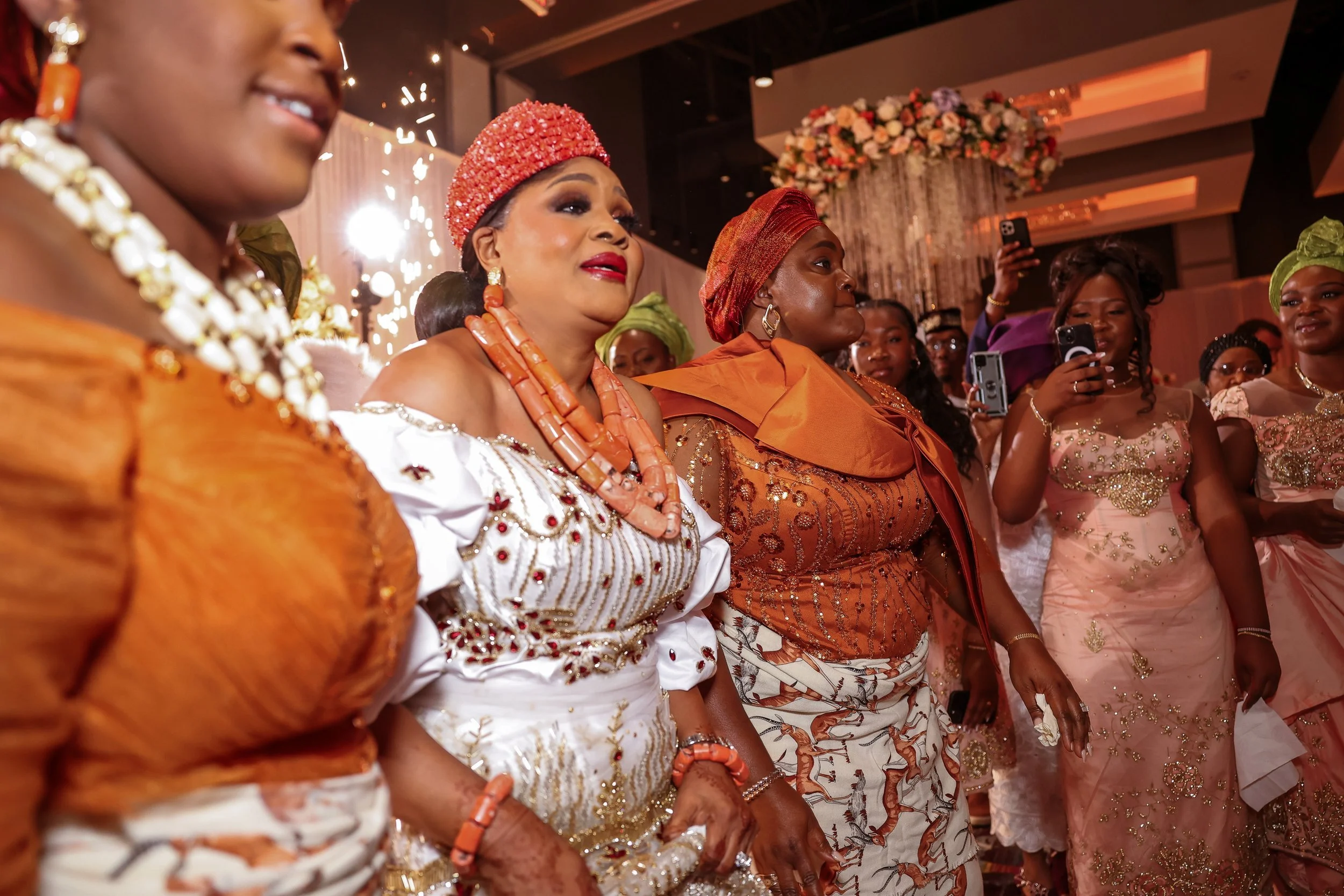 Group of women dressed in traditional Nigerian attire at a celebration or cultural event, with colorful clothing, jewelry, and headpieces, in an elegant decorated venue.