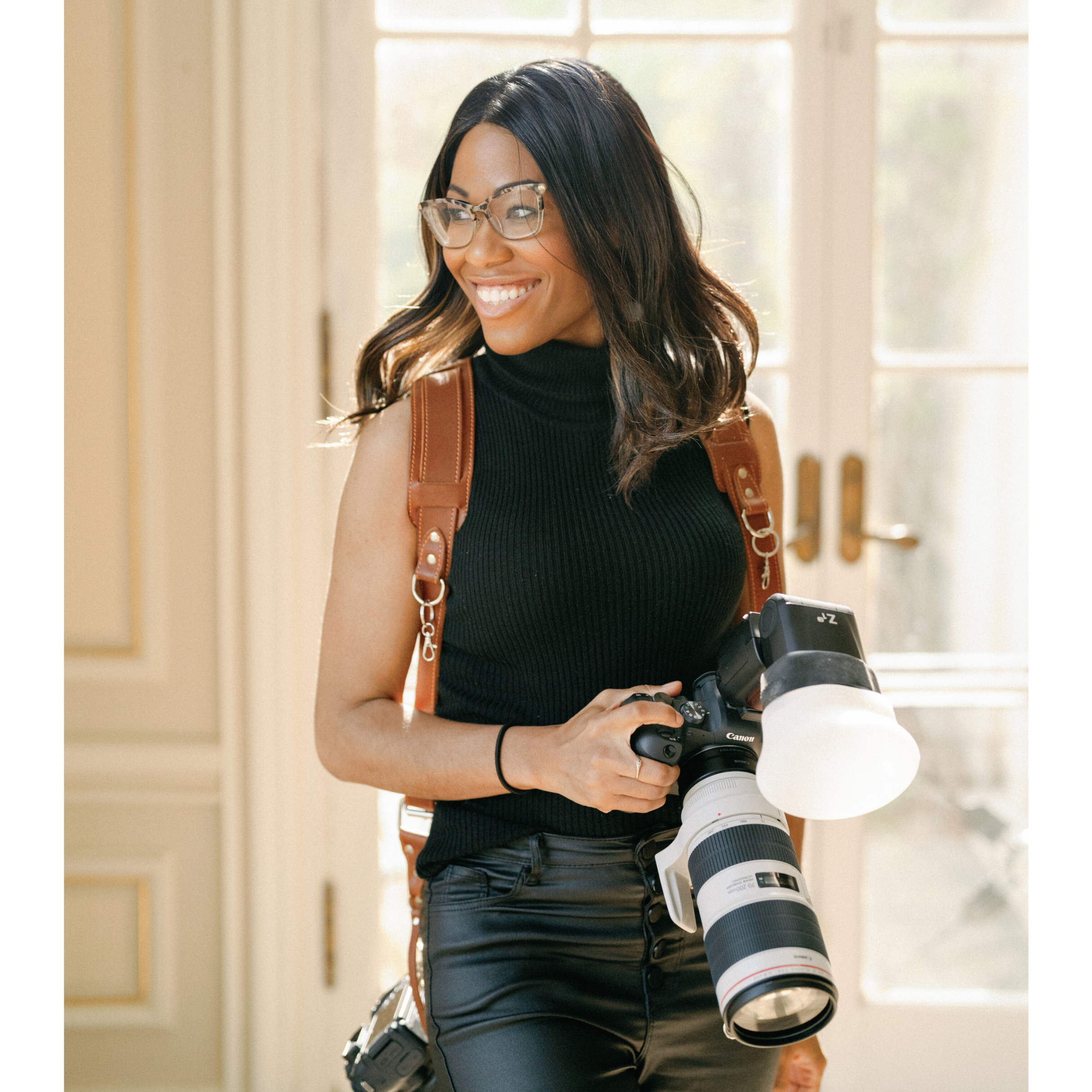 A woman with glasses wearing a black sleeveless turtleneck, carrying a brown leather backpack, holding a professional camera with a large lens and a flash attachment, smiling and standing indoors near large windows.