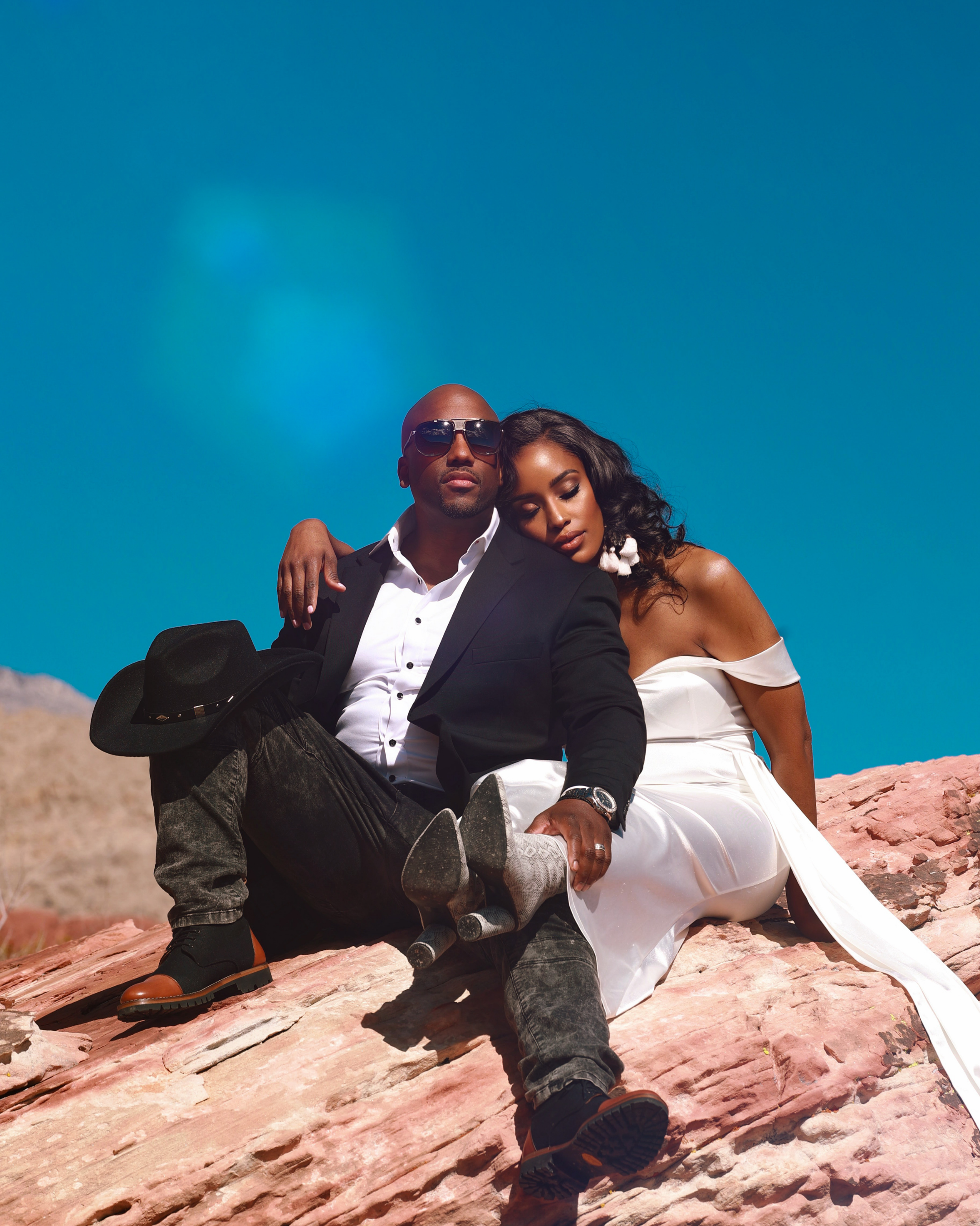 A man and woman sitting together on pink rocks in a desert landscape under a blue sky. The man wears sunglasses, a black suit jacket, white shirt, dark jeans, and brown boots, with a black hat resting on his knee. The woman wears a white off-shoulder