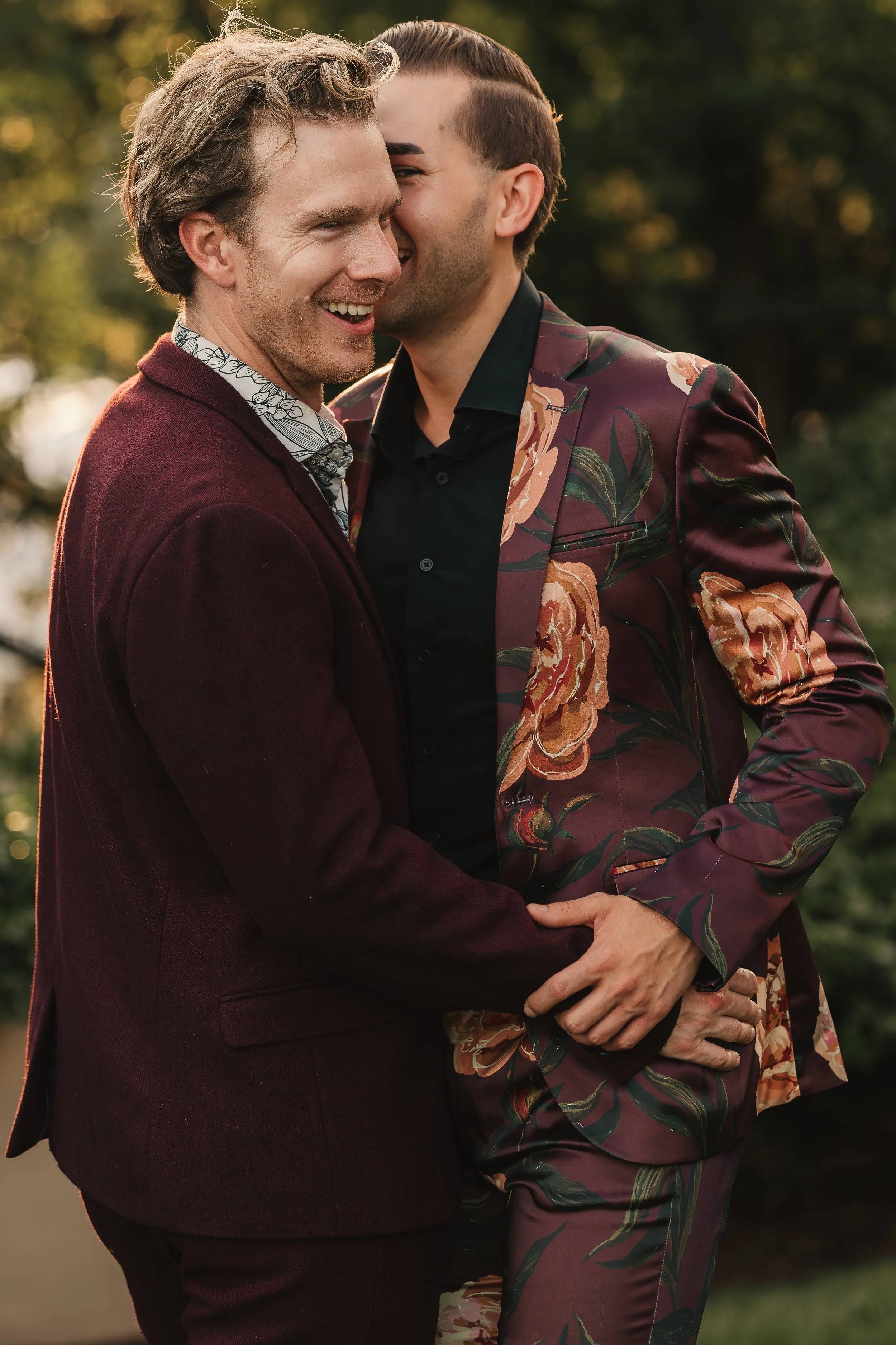 Two men are smiling and holding hands while standing outside during sunset, dressed in elegant clothing, with trees in the background.