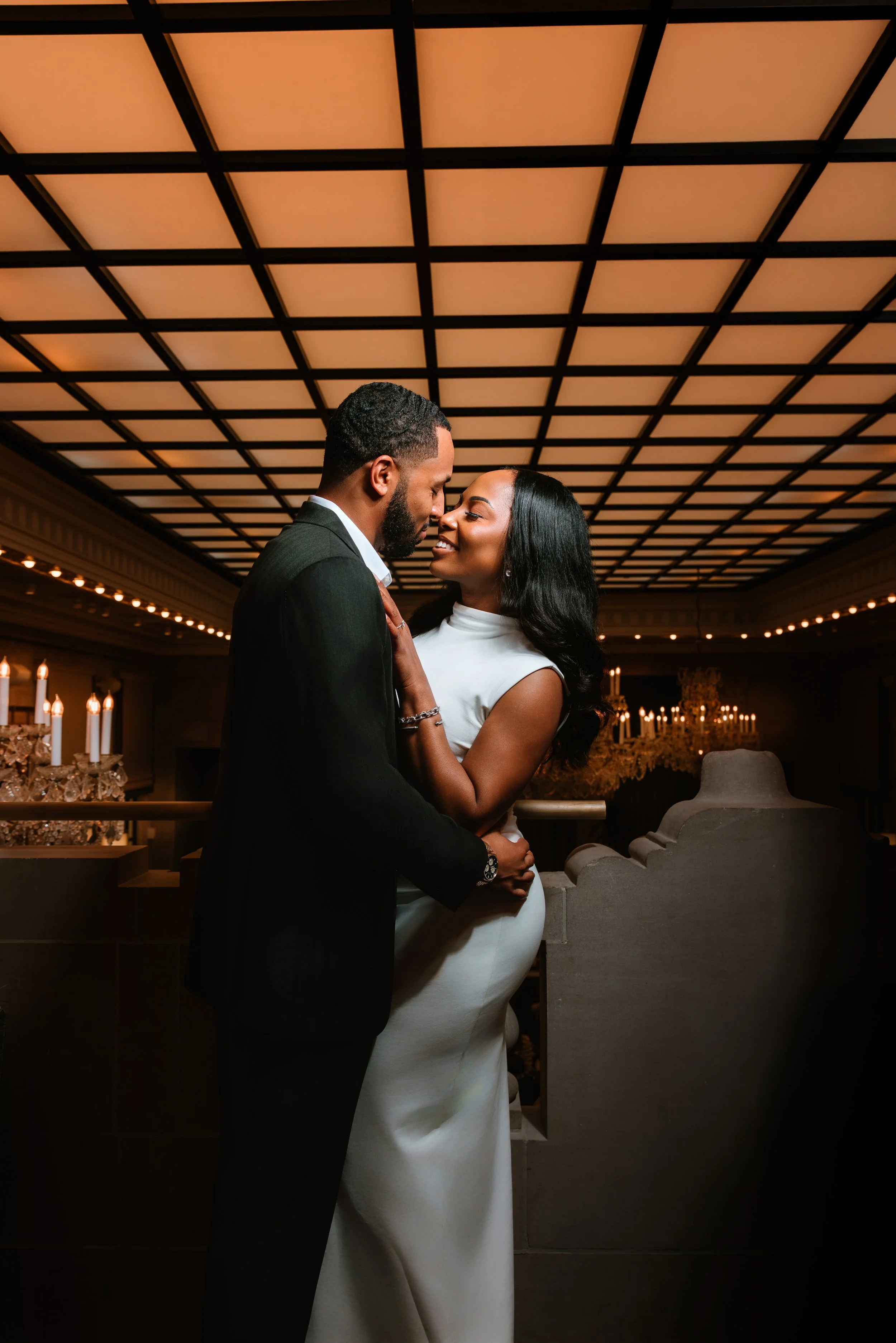A romantic couple stands close together in an elegant indoor setting with warm lighting, looking into each other's eyes. The woman is wearing a white dress, and the man is dressed in a black suit. The ceiling has a geometric grid pattern, and there's