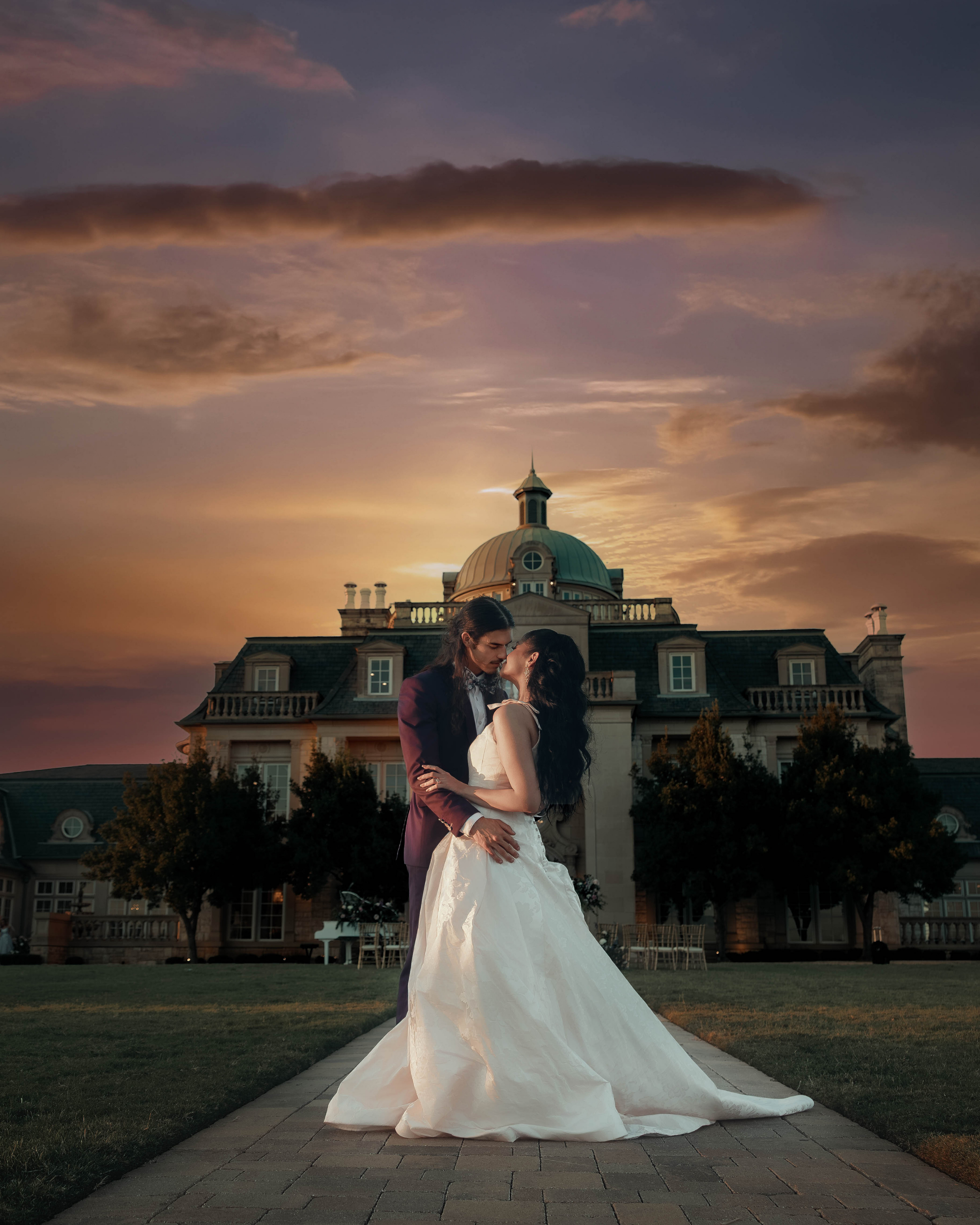 Couple dressed in wedding attire sharing a romantic moment at sunset front of a grand mansion.