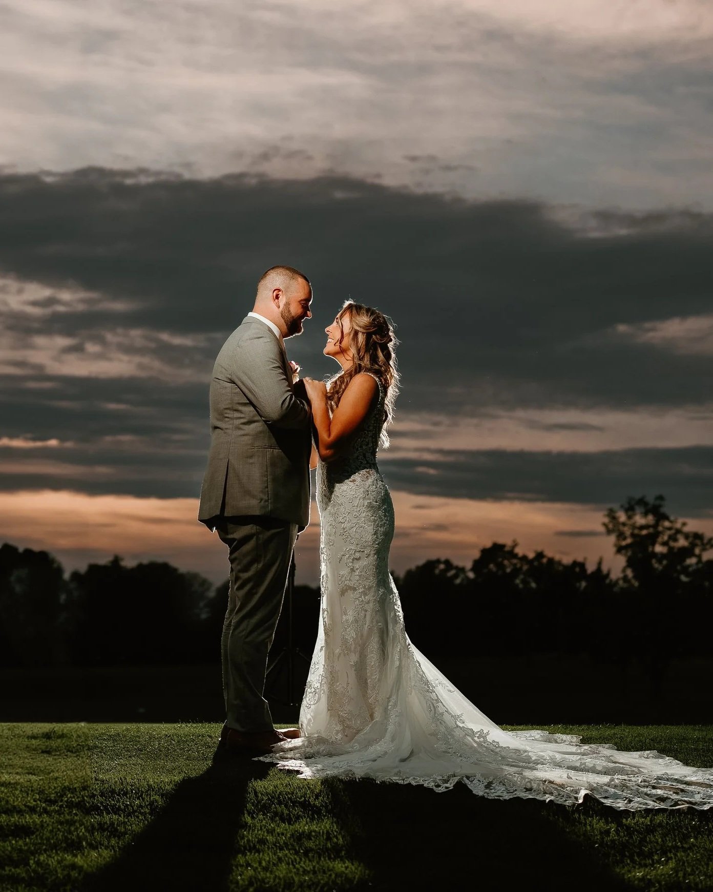 A bride and groom standing close together outdoors at sunset, smiling and looking at each other, with the bride in a white lace wedding gown and the groom in a gray suit.