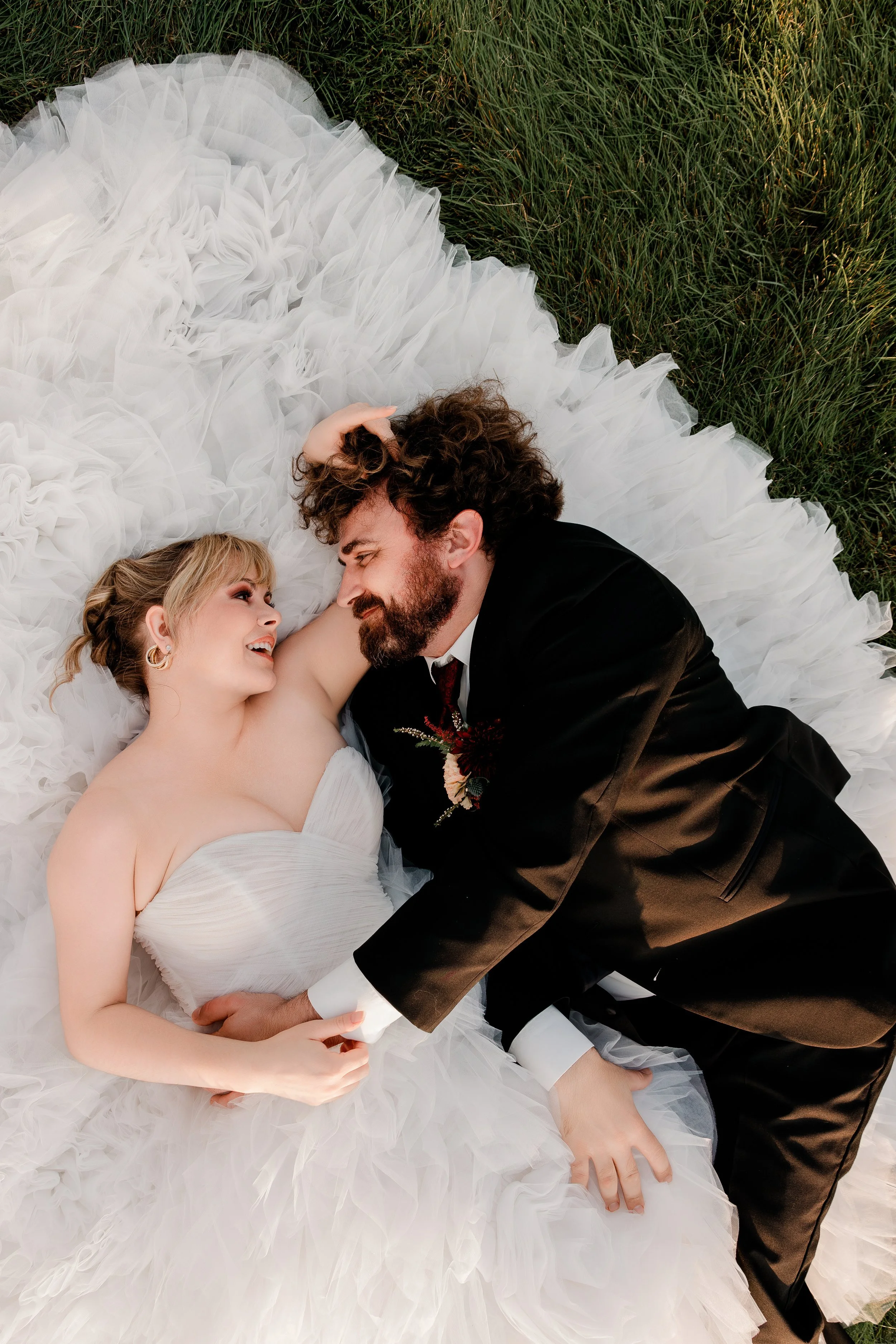 A couple in wedding attire lying on a white, ruffled fabric on grass, smiling at each other.