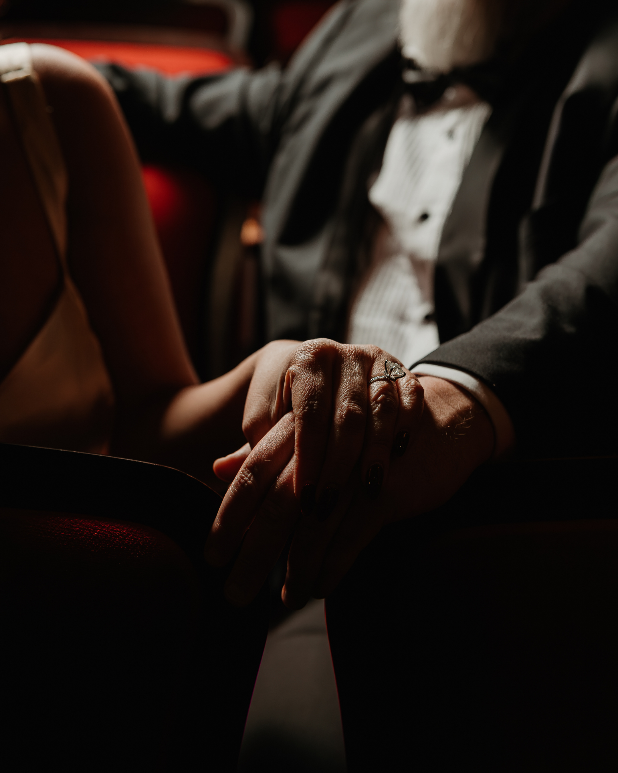 Close-up of a man and woman holding hands, with the woman's hand resting on top of the man's hand, in a dimly lit setting.