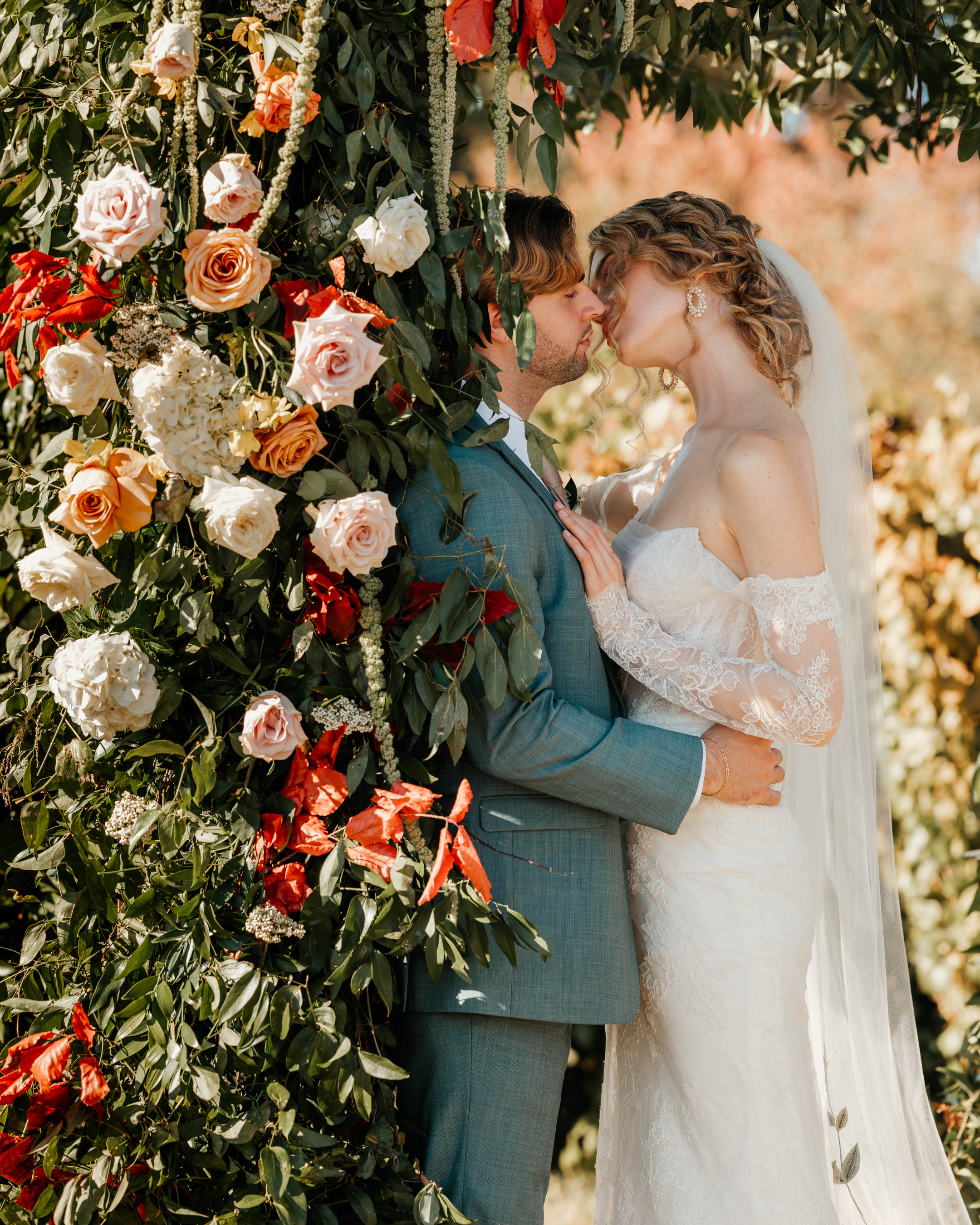 A bride and groom stand close with foreheads touching, surrounded by a floral wedding arch of roses and greenery outdoors.