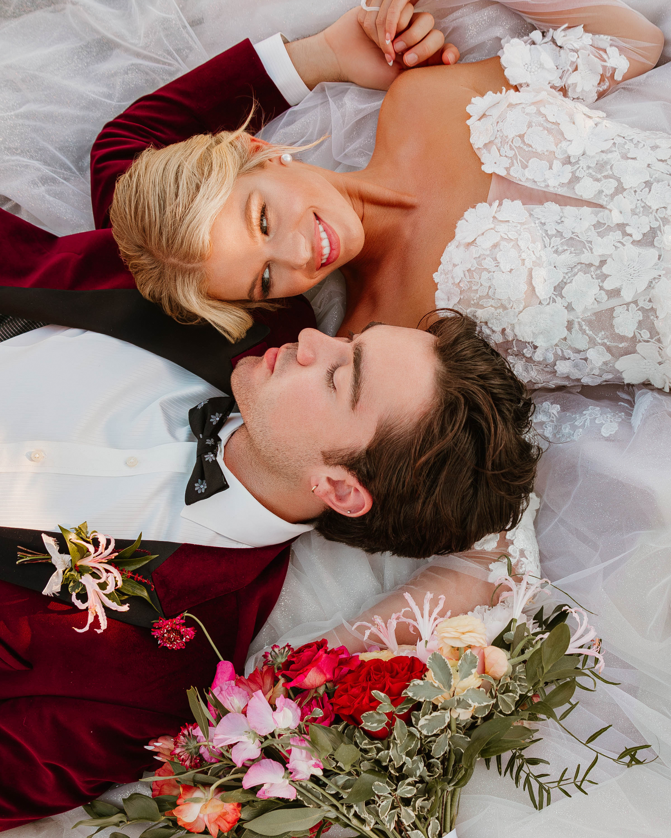 Bride and groom lying on a bed, facing each other, with the bride smiling and holding a bouquet of flowers.