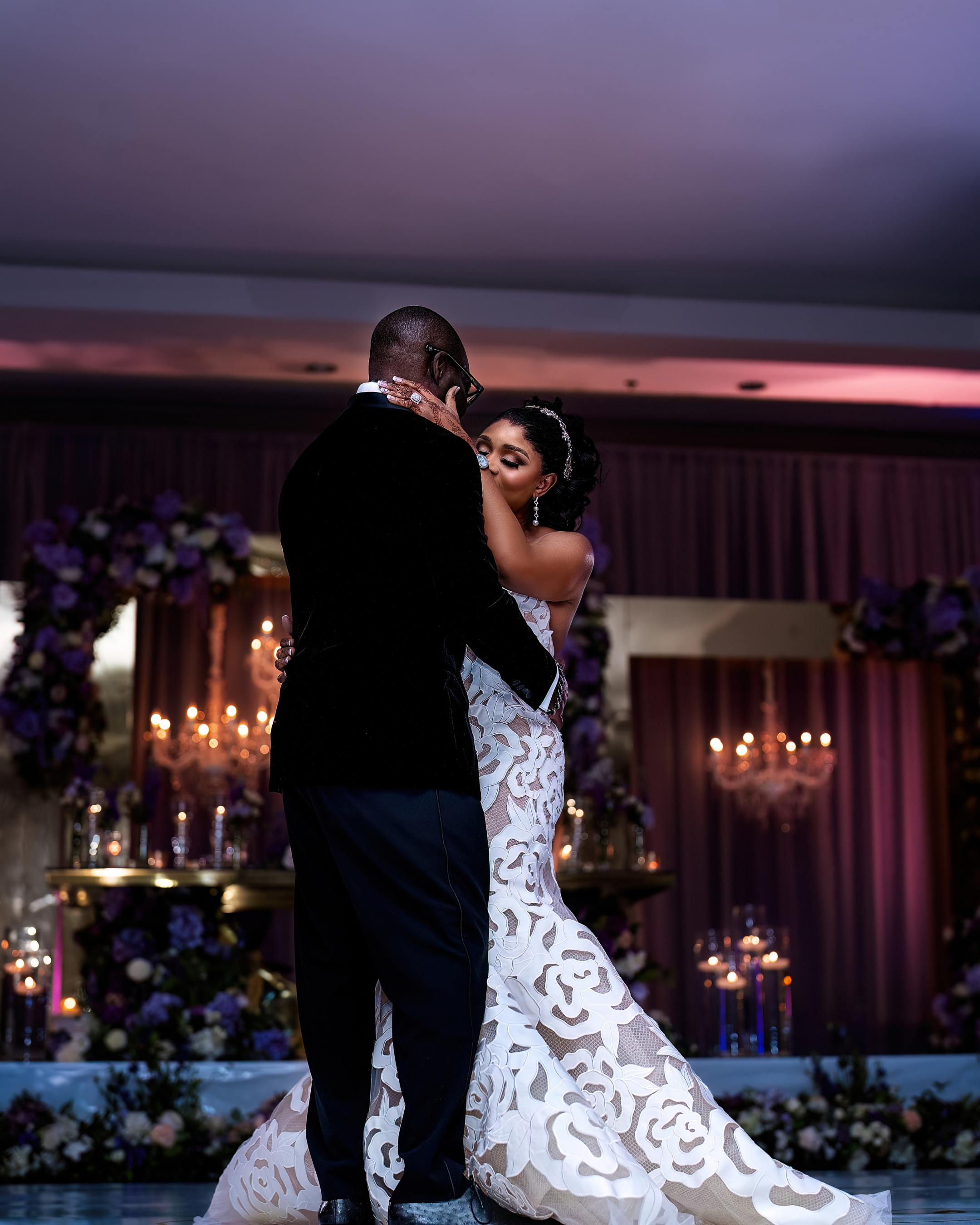 A bride and groom sharing their first dance at a wedding reception, with purple and white floral decorations and candles in the background.