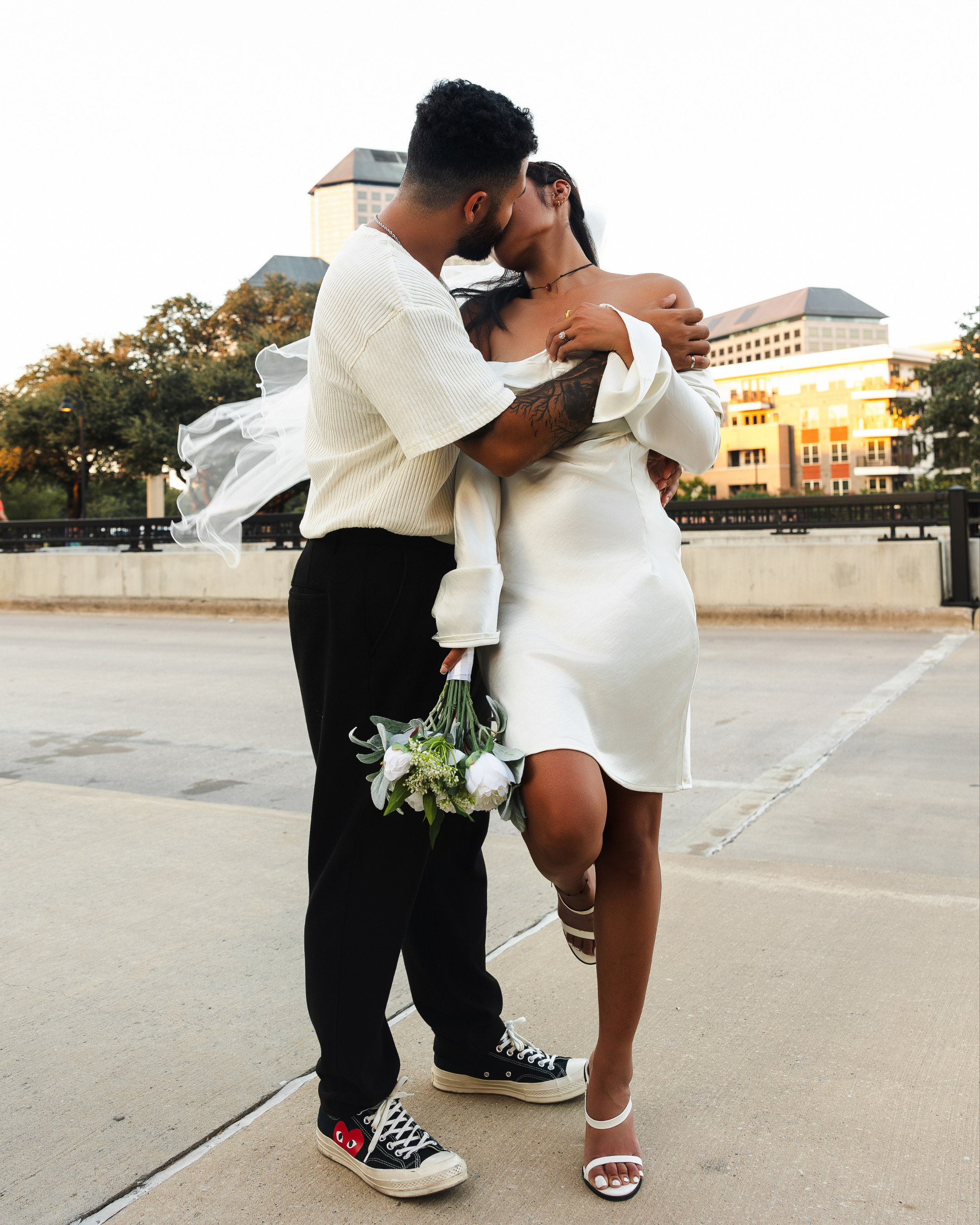 A couple kisses on a city street during sunset, woman holding flowers, dressed in white, man in casual clothes and sneakers.