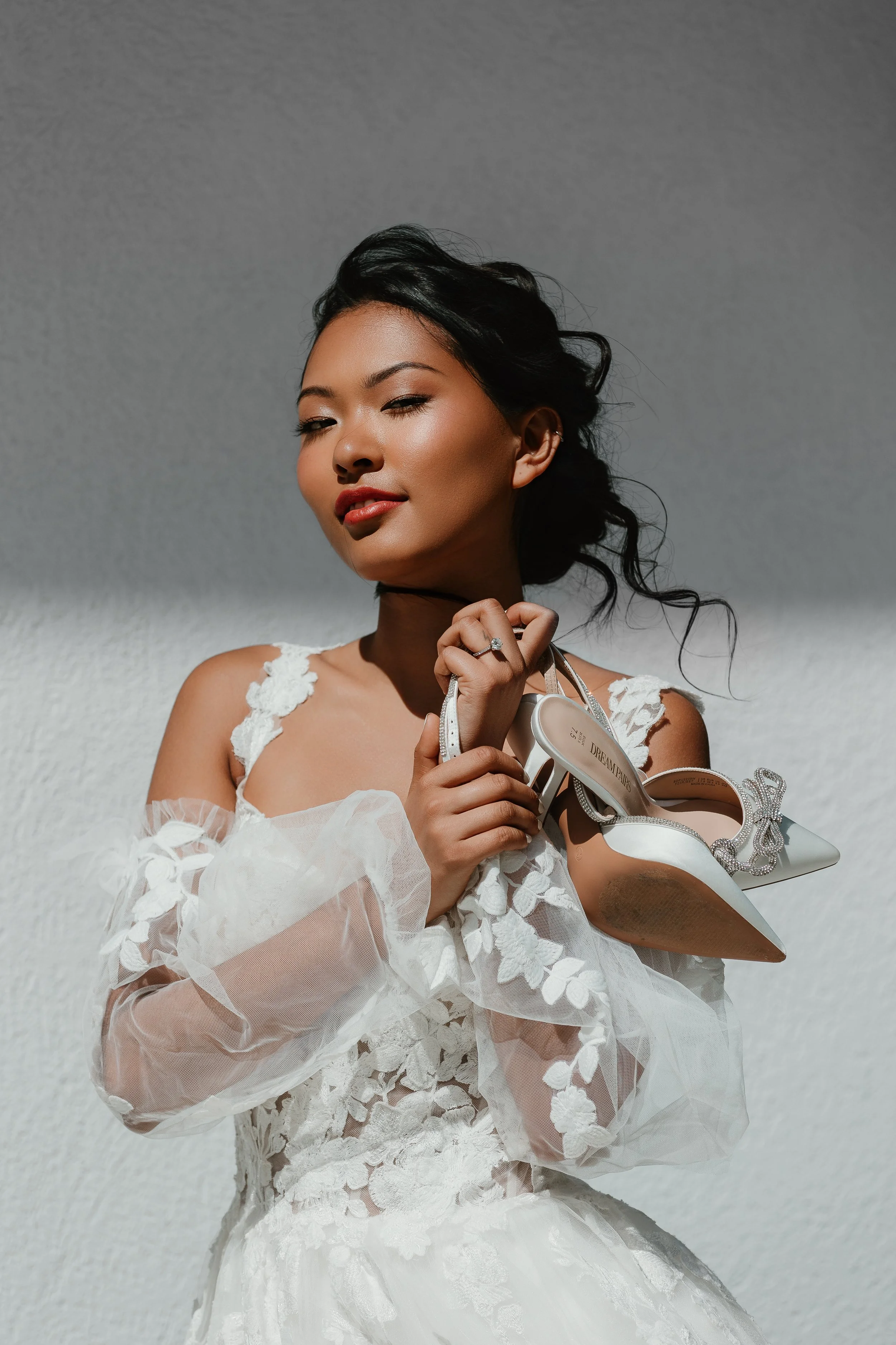 A woman in a white lace dress holding a pair of white high heels with embellishments.