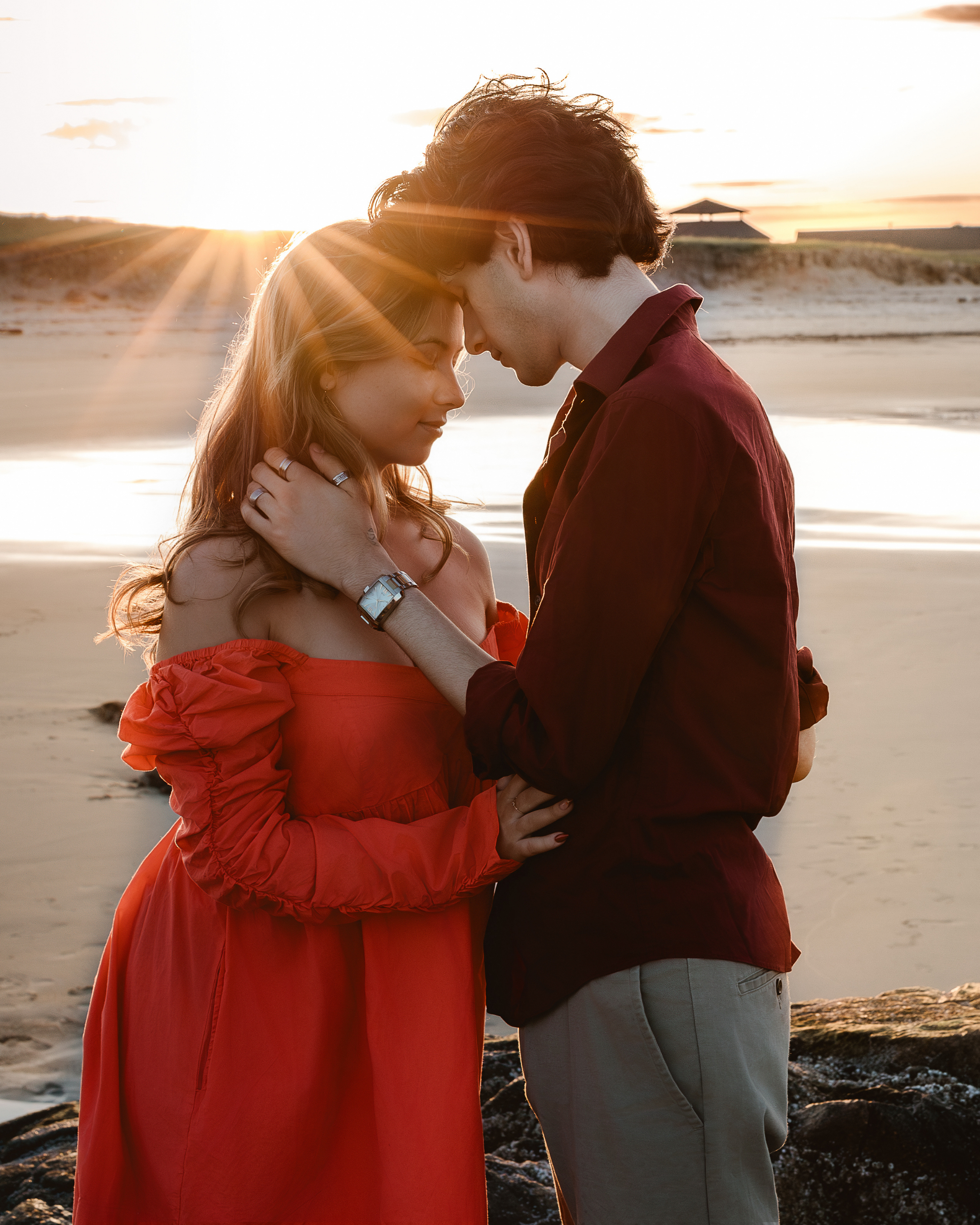 A romantic couple standing close together on a beach during sunset, touching foreheads and holding each other.