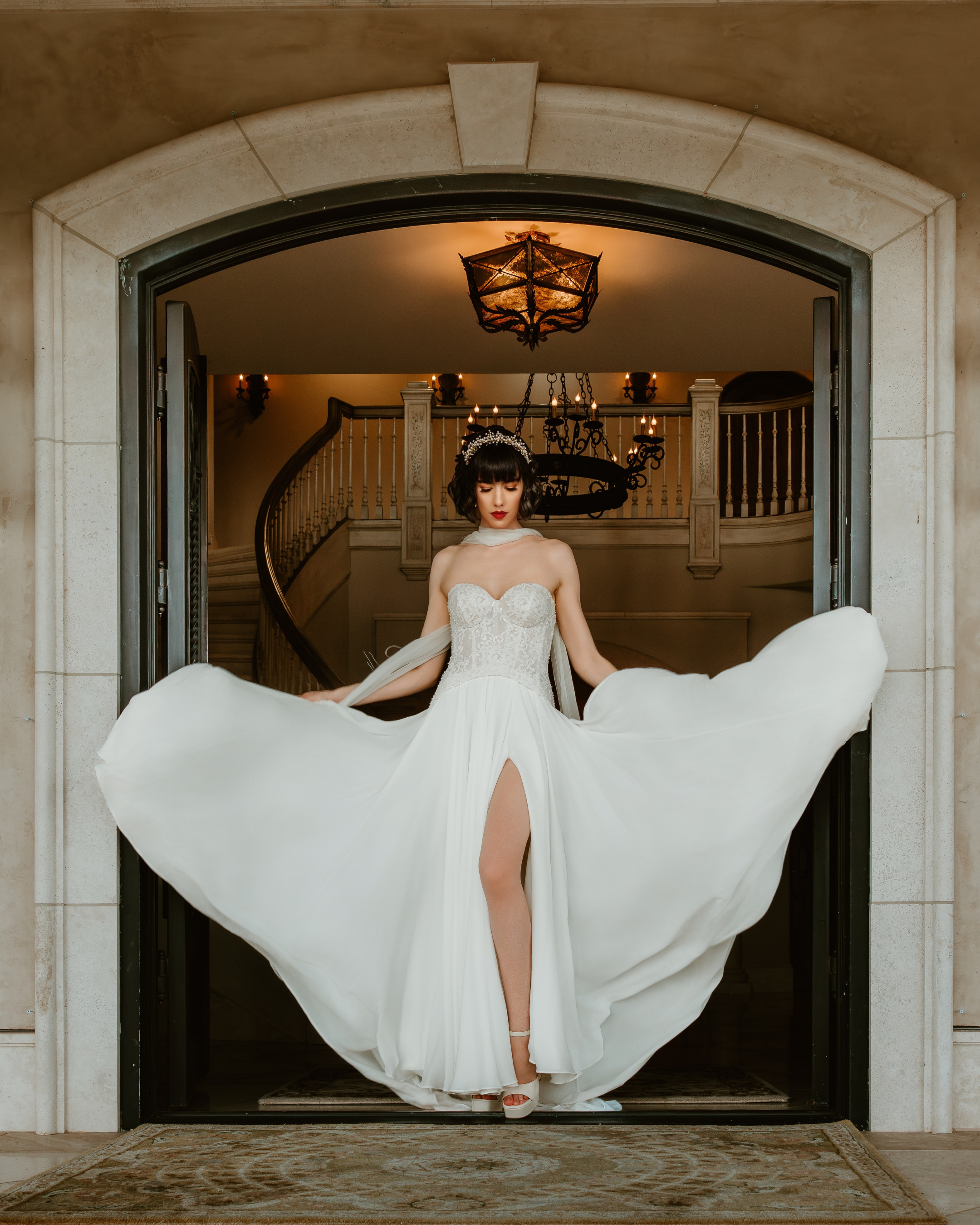 A woman in a white wedding dress with a slit, standing in an open doorway inside a grand house with a staircase and chandelier in the background.