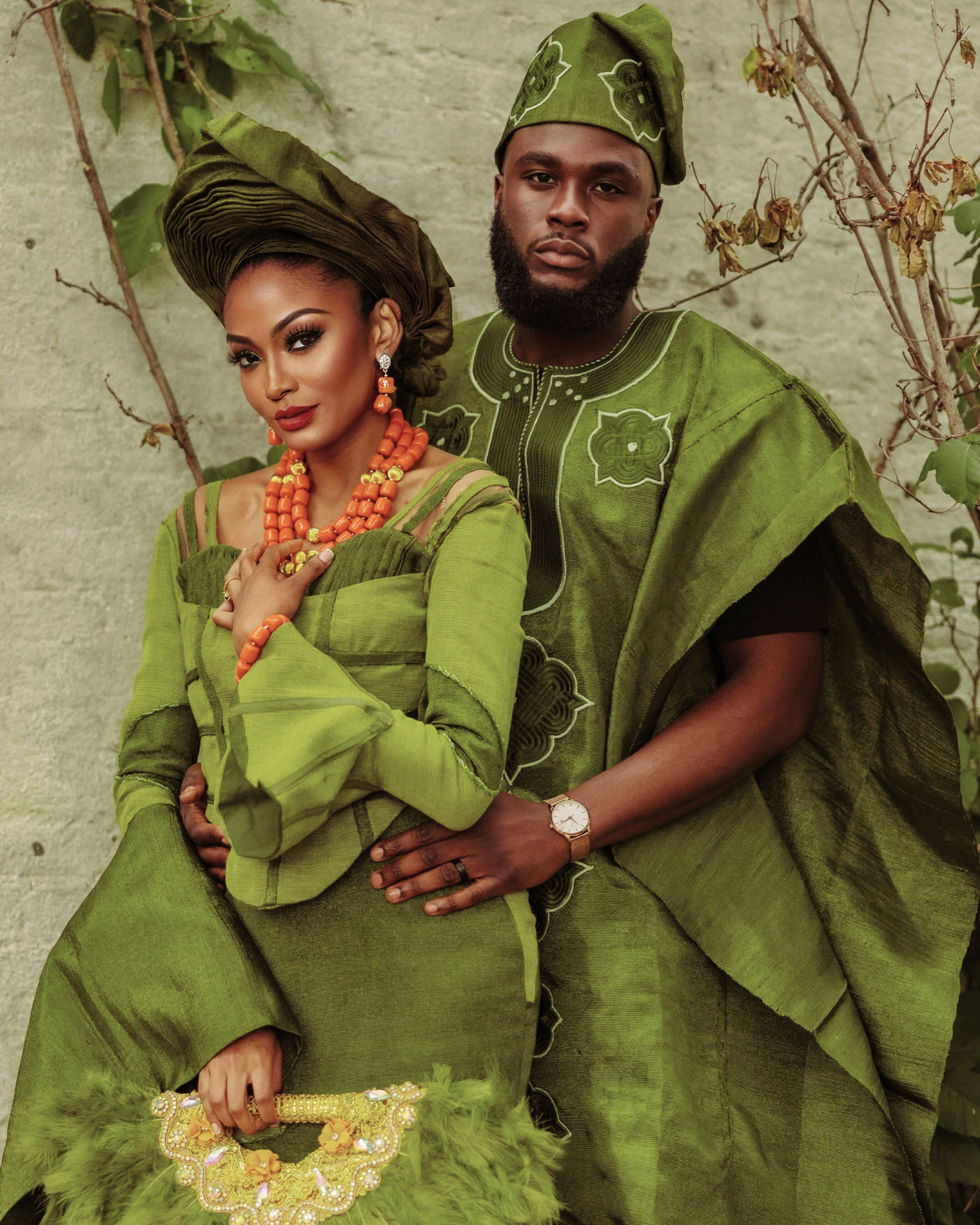 A man and woman dressed in traditional green African attire, posing together outdoors against a textured wall with dried vines. The woman wears elaborate headwrap, jewelry, and a gown with feathered embellishments, while the man wears a matching outf