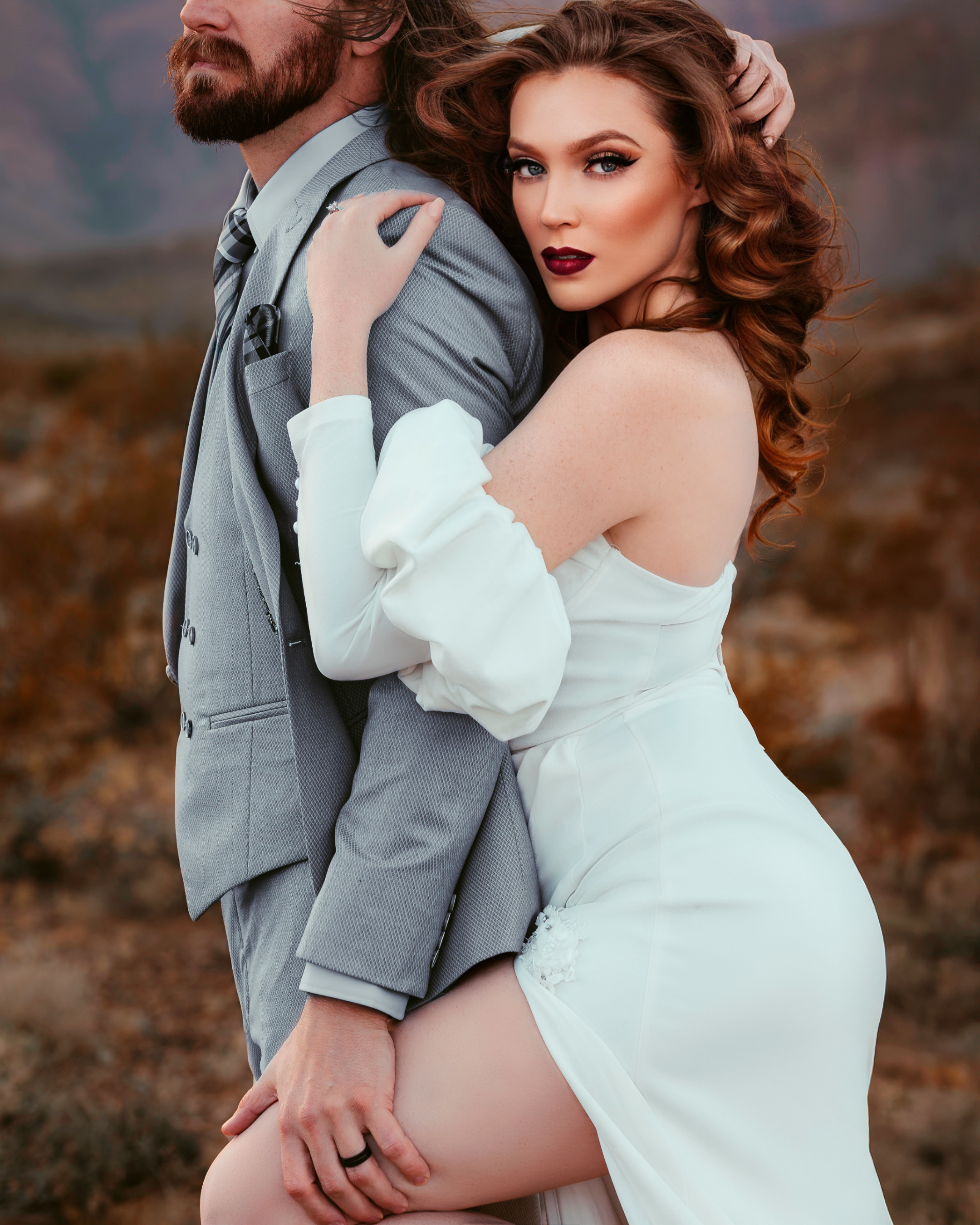 A woman with red hair and makeup in a white dress hugging a man with a beard in a gray suit outdoors.