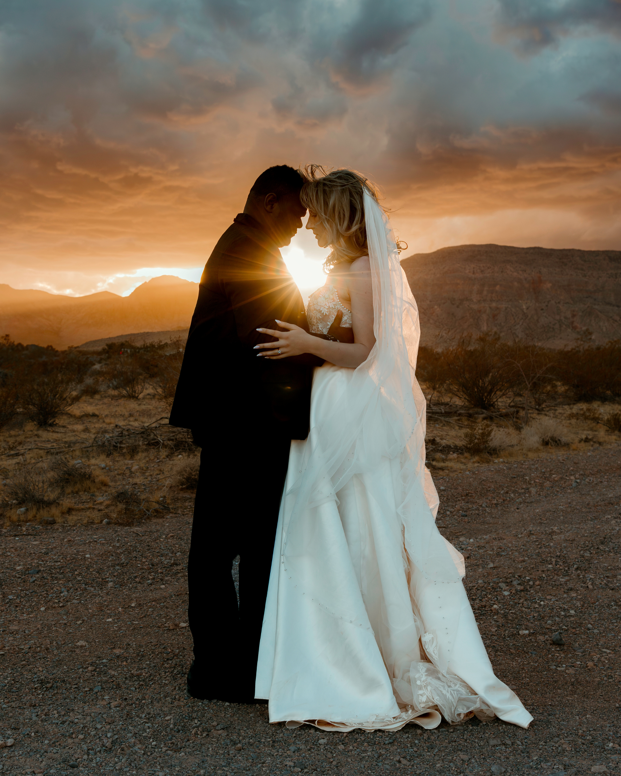 A bride and groom embracing at sunset in a desert landscape with mountains and cloudy sky.