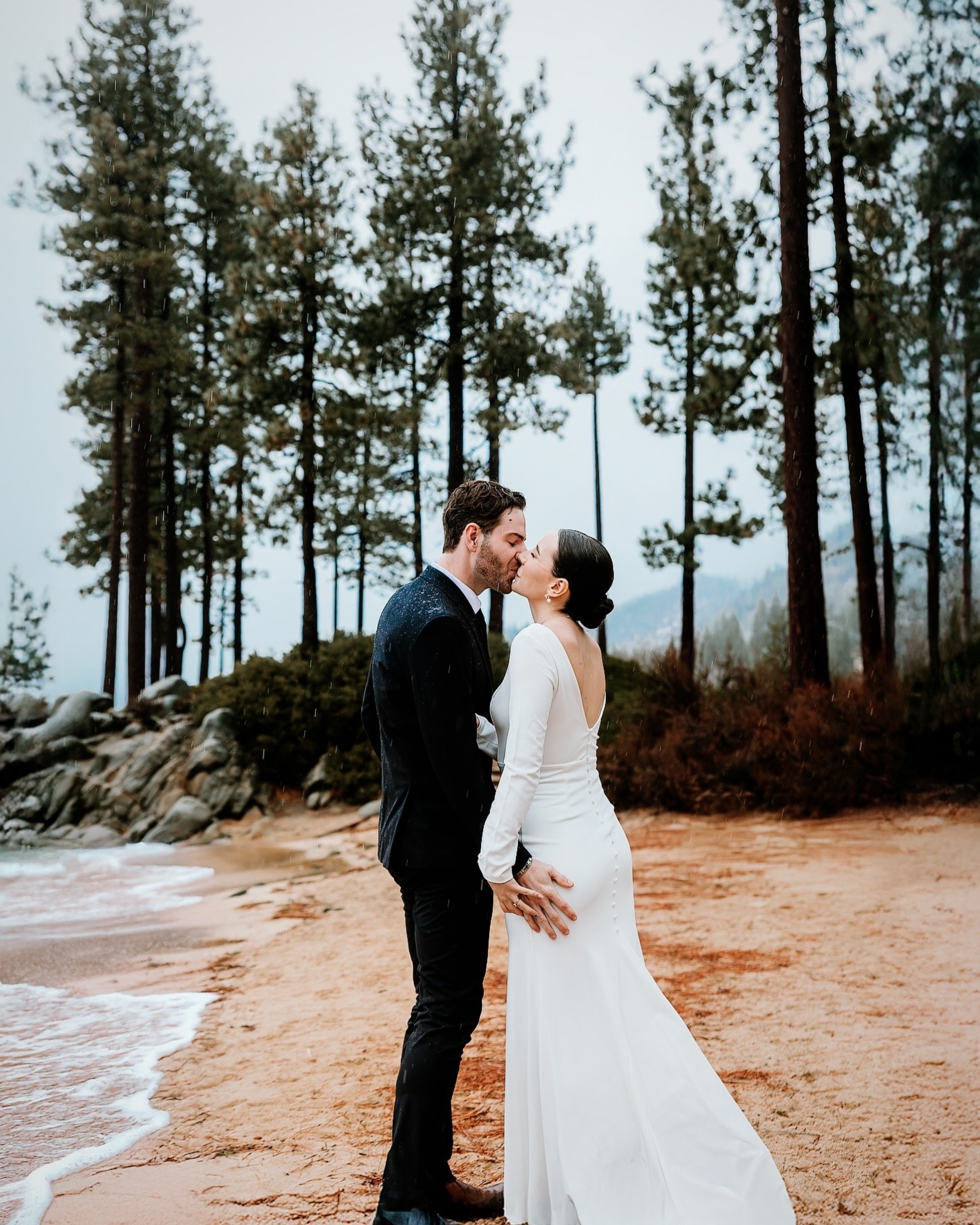 A couple in wedding attire sharing a kiss on a beach with tall pine trees in the background.