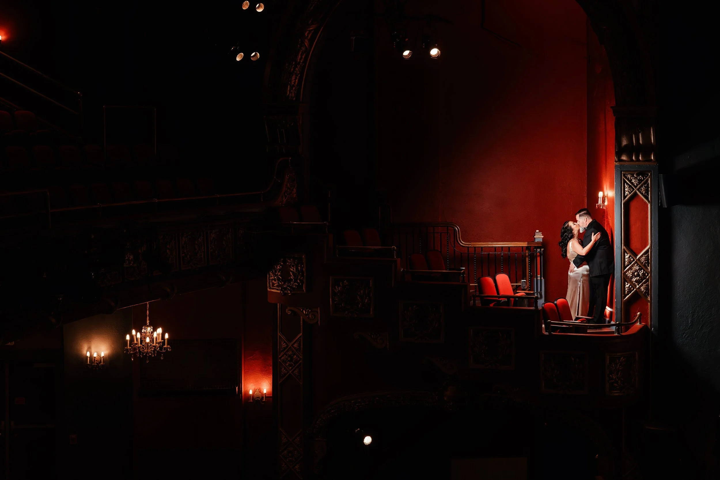 A couple is kissing in a theater box with red walls, surrounded by empty red chairs, dim lighting, chandelier, and wall sconces.