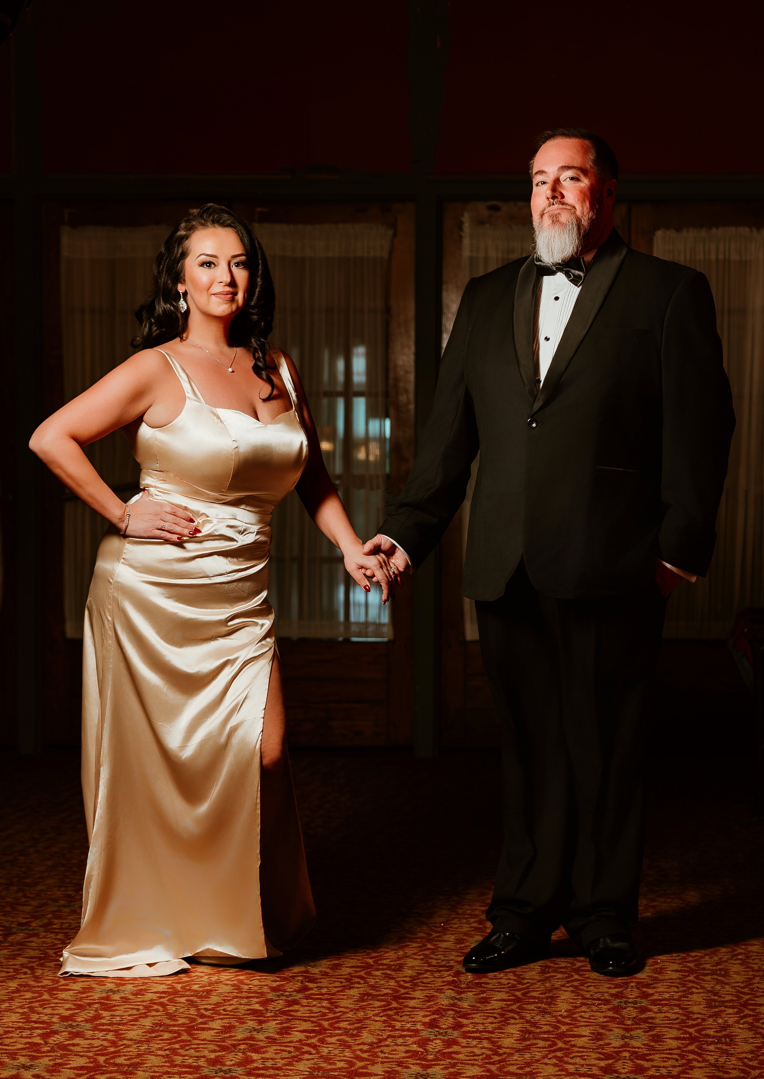 A man and woman dressed in formal attire, holding hands in a dimly lit room with curtains and wooden panels.