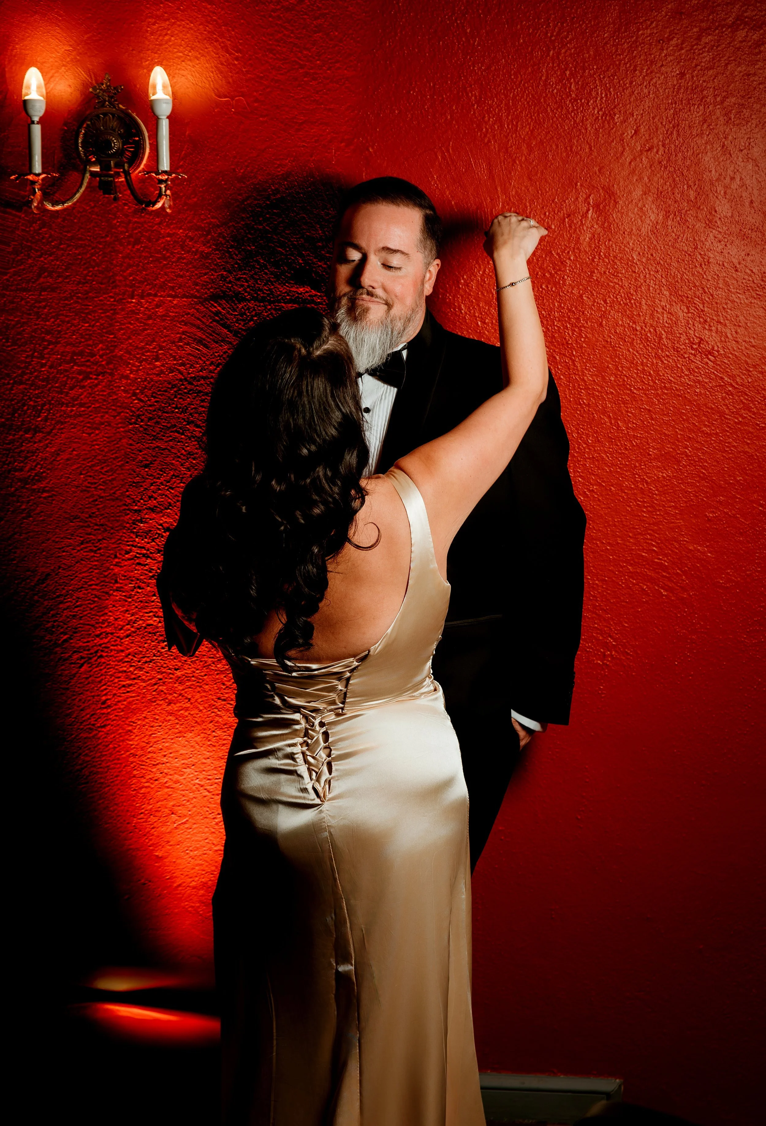 A woman in a satin champagne-colored dress with lace-up back dancing with an older man in a tuxedo in front of a red wall with a candle wall sconce.