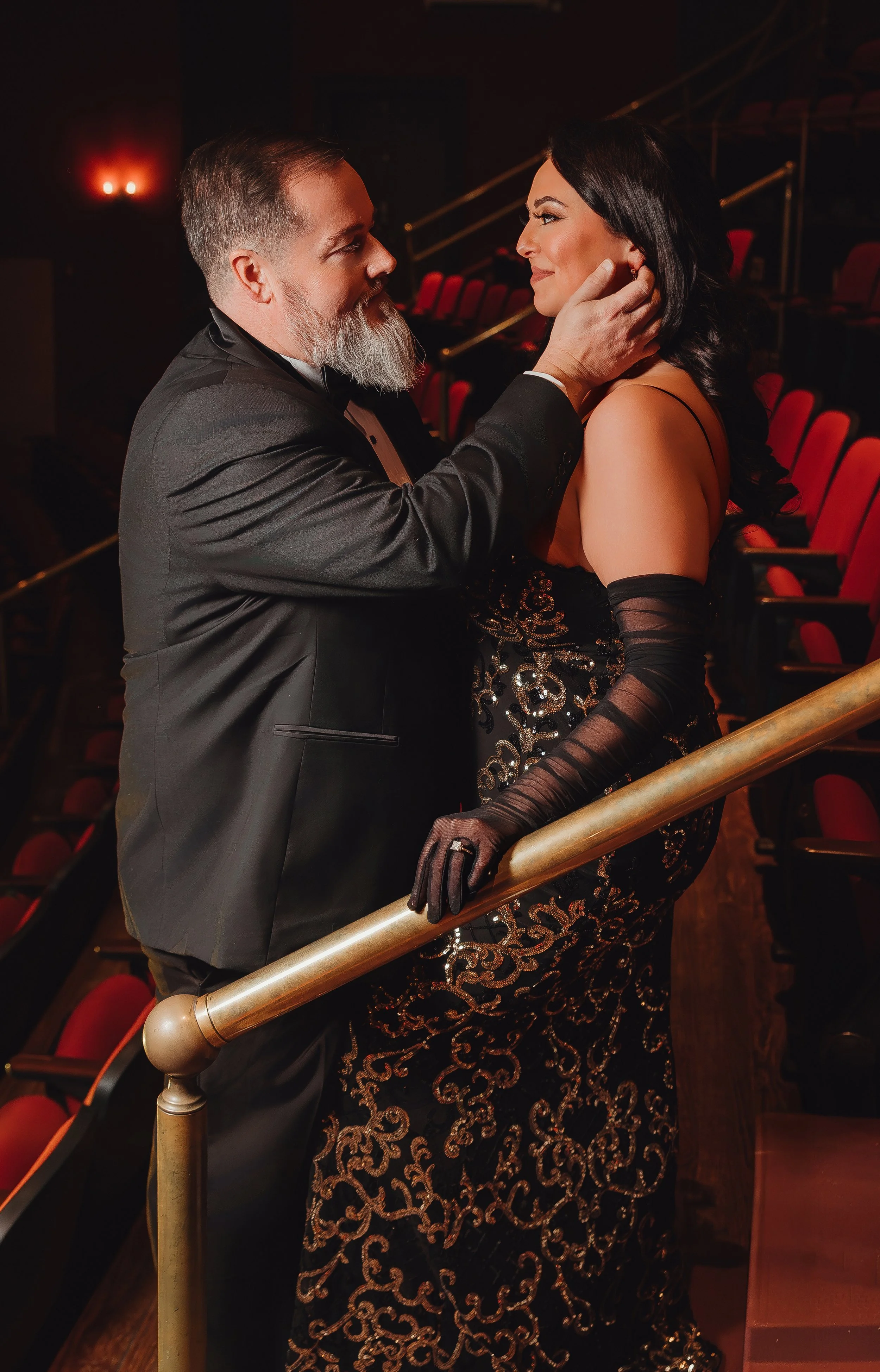 A man in a tuxedo and a woman in an elegant floral gown and gloves share an intimate moment backstage at a theater, with empty red seats in the background.