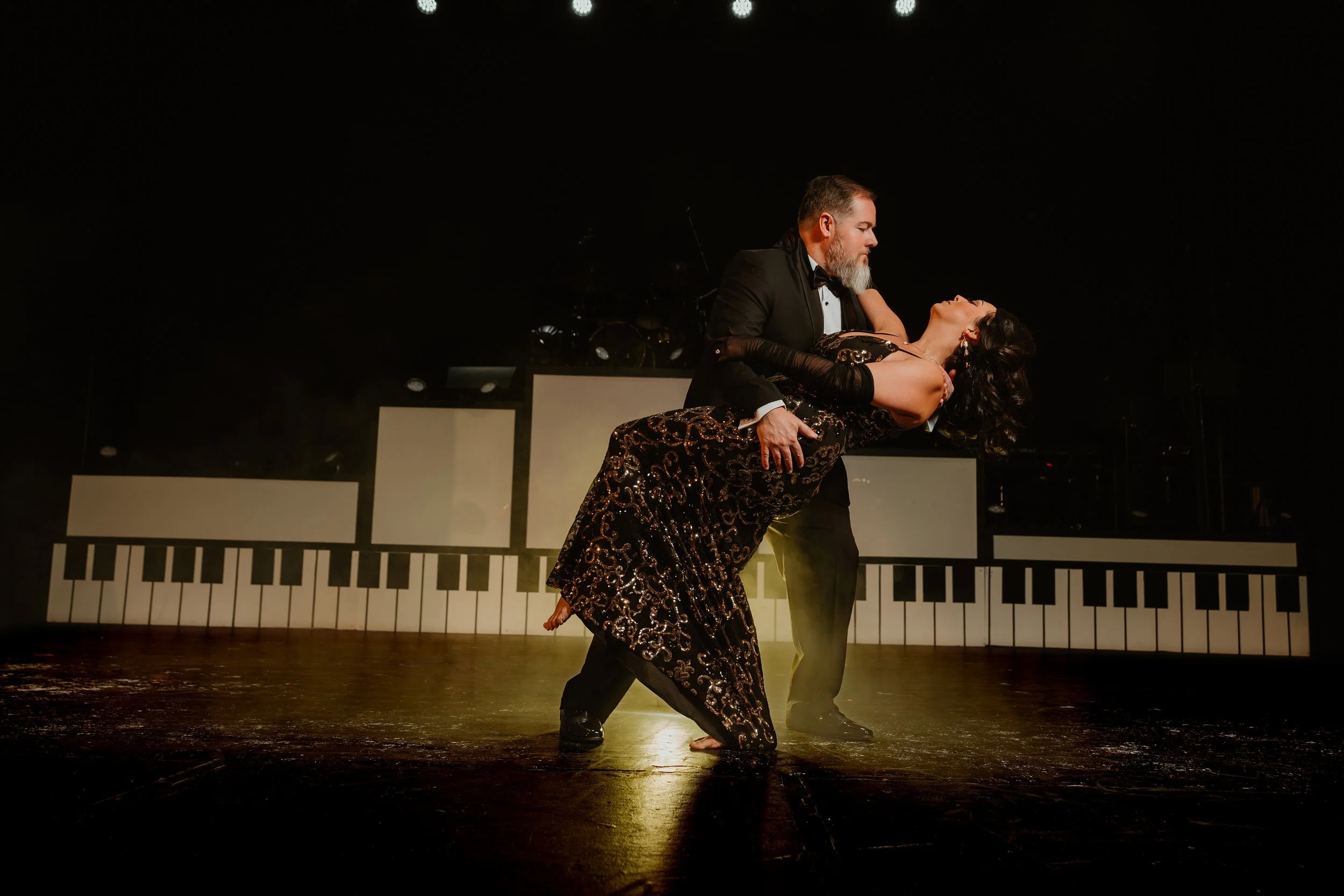 A man and woman perform a dance on stage with a piano keyboard backdrop. The man, dressed in a black tuxedo, holds the woman, who is wearing a black and gold sequined dress, in a dramatic dance pose.