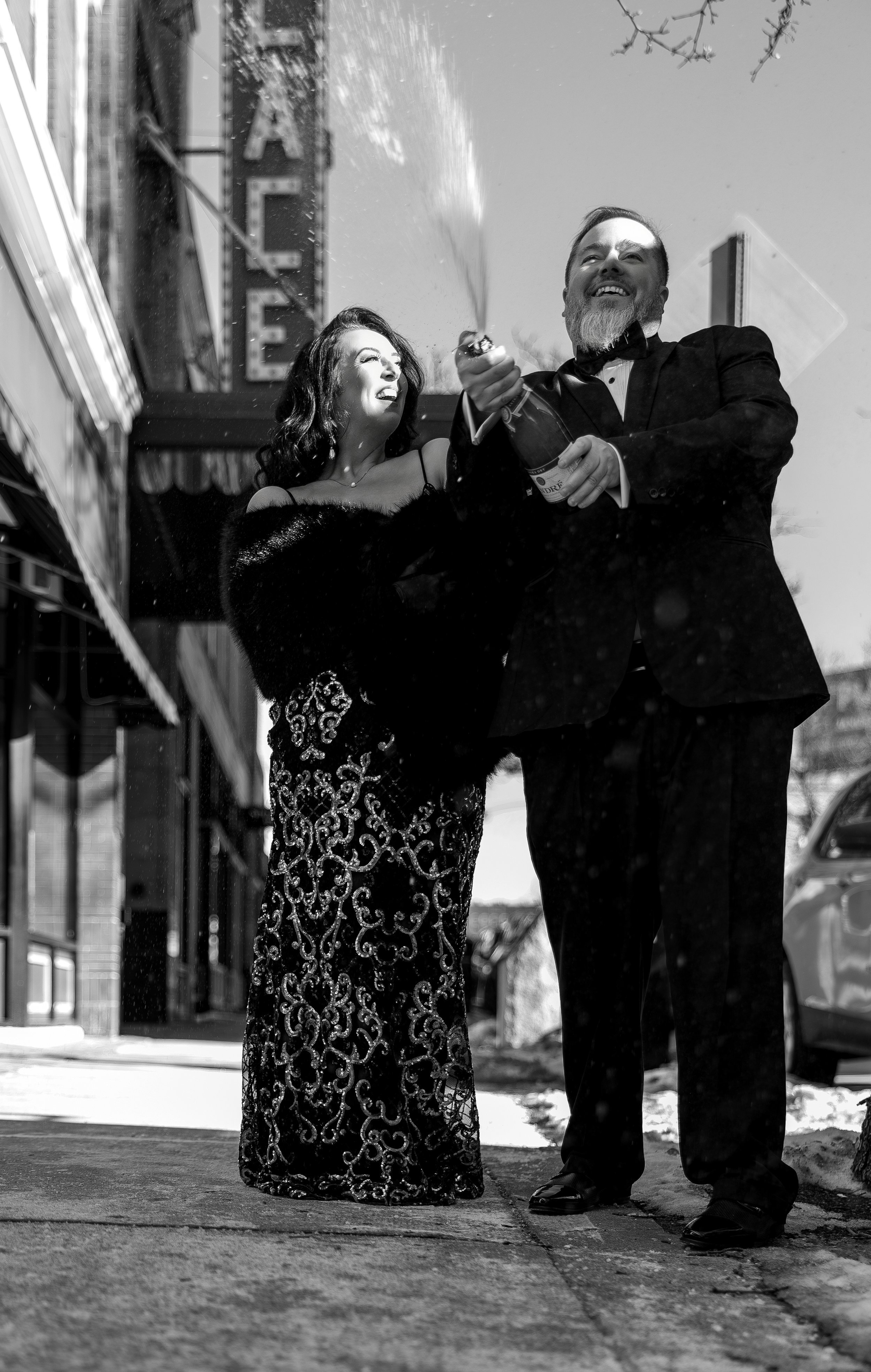 A black and white photo of a man and woman celebrating on a city sidewalk, opening a bottle of champagne with water spraying out, both smiling and dressed formally.
