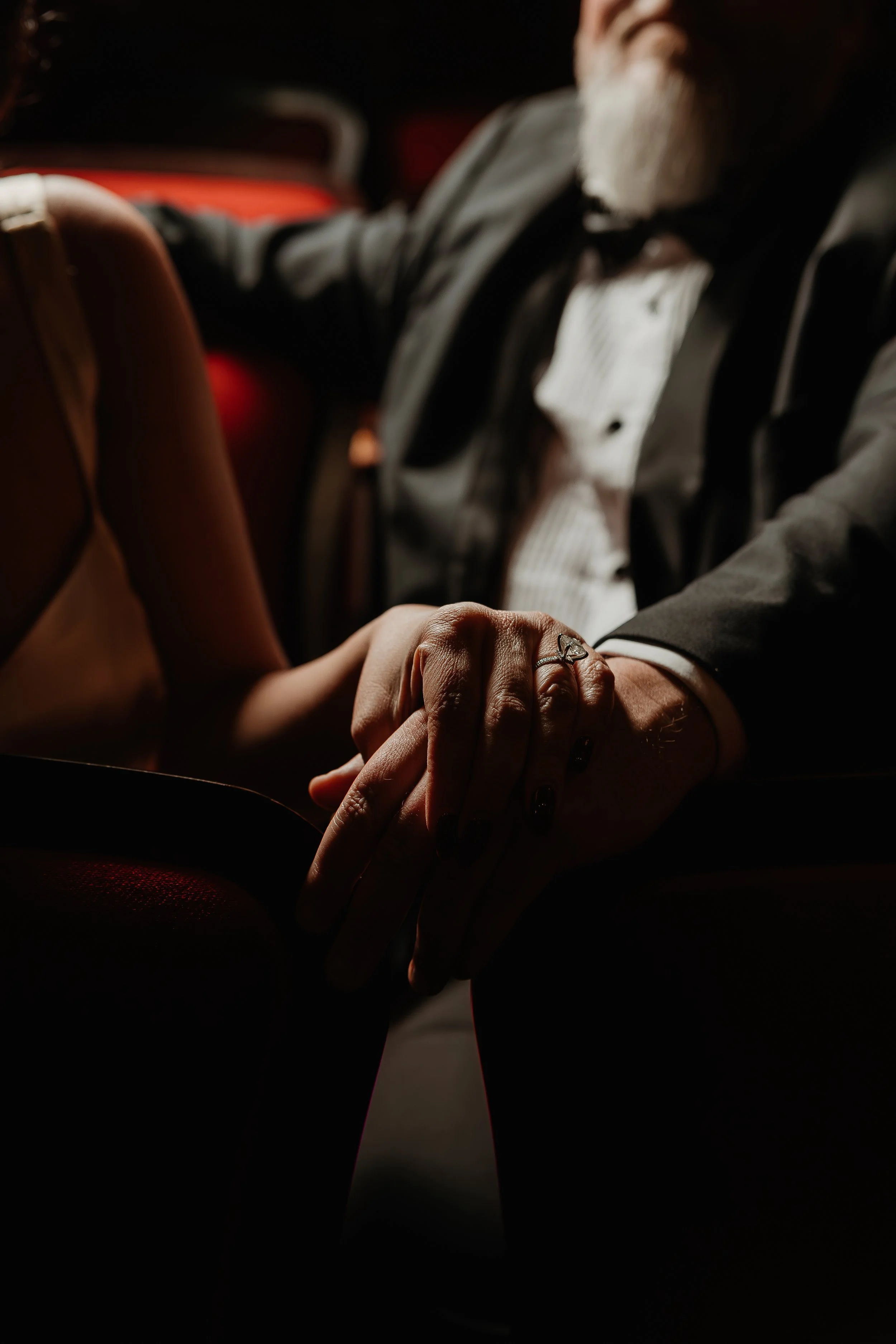 A couple holding hands at a formal event, with a woman wearing a spaghetti strap dress and a man in a tuxedo.