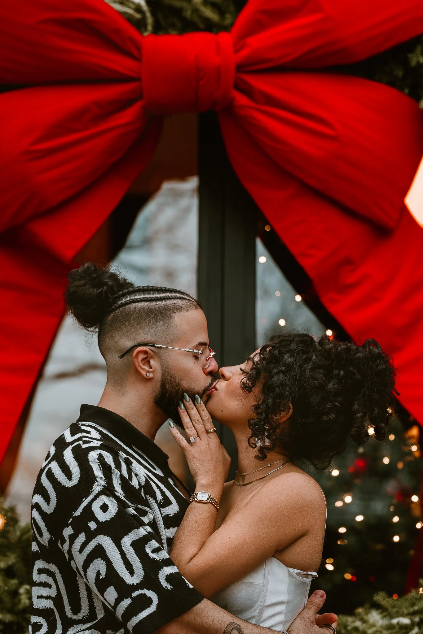 A couple sharing a kiss under a large red bow with Christmas decorations in the background.