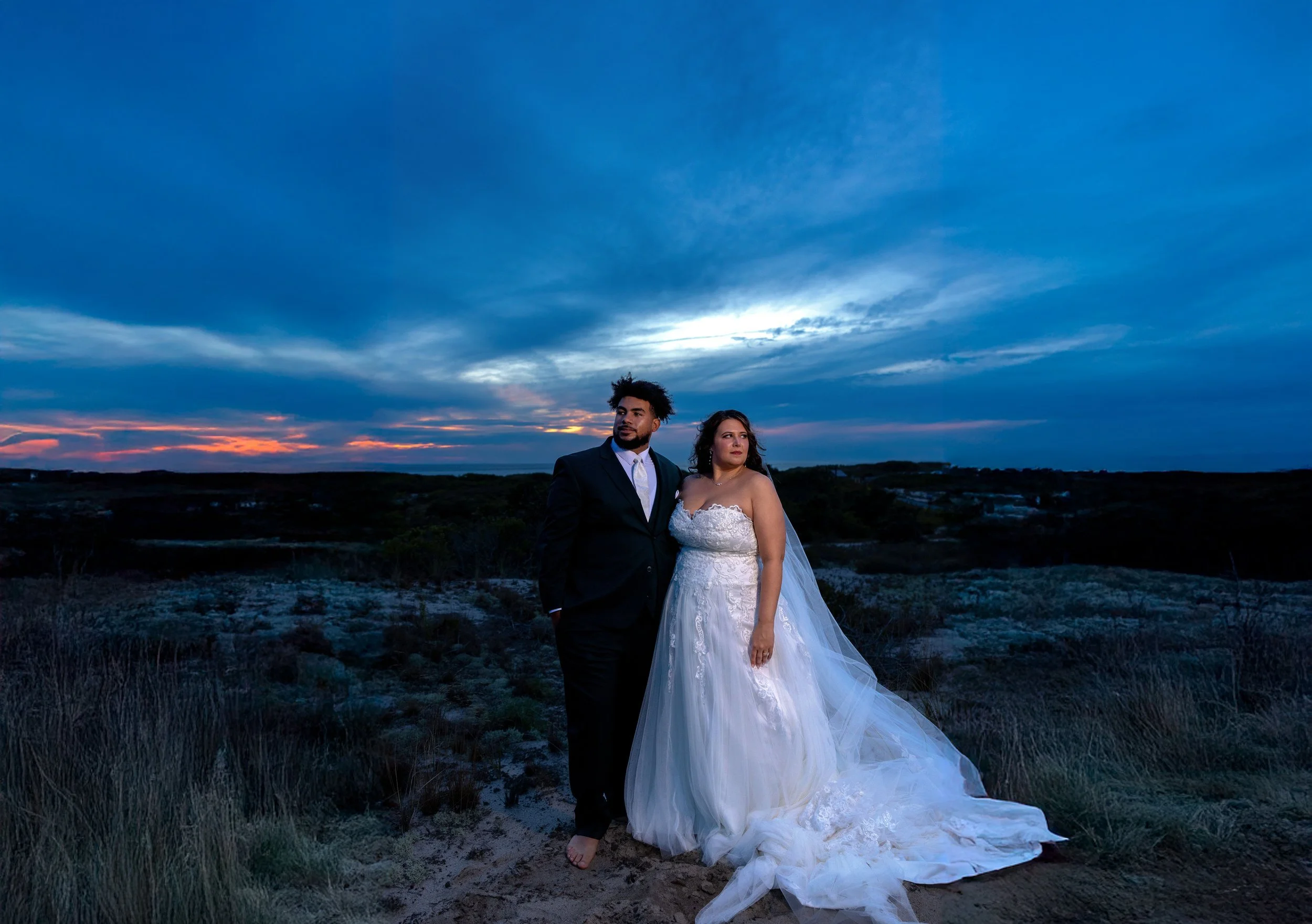 A couple dressed in wedding attire standing outdoors at sunset, with a dramatic blue sky and horizon in the background.