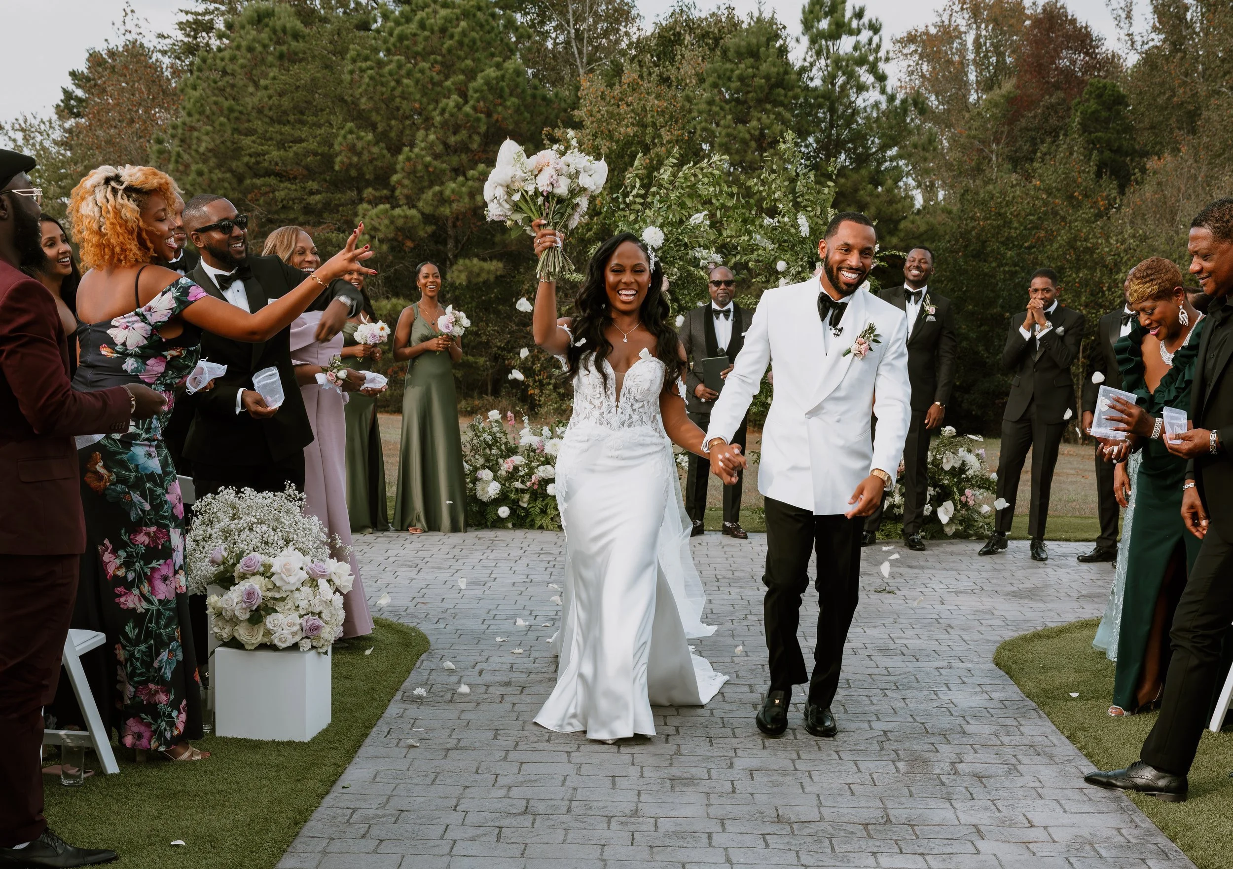 Bride and groom walking down an outdoor aisle, holding hands, smiling; bride raising a bouquet in celebration; guests dressed in formal attire, cheering and clapping; floral arrangements and greenery in the background.