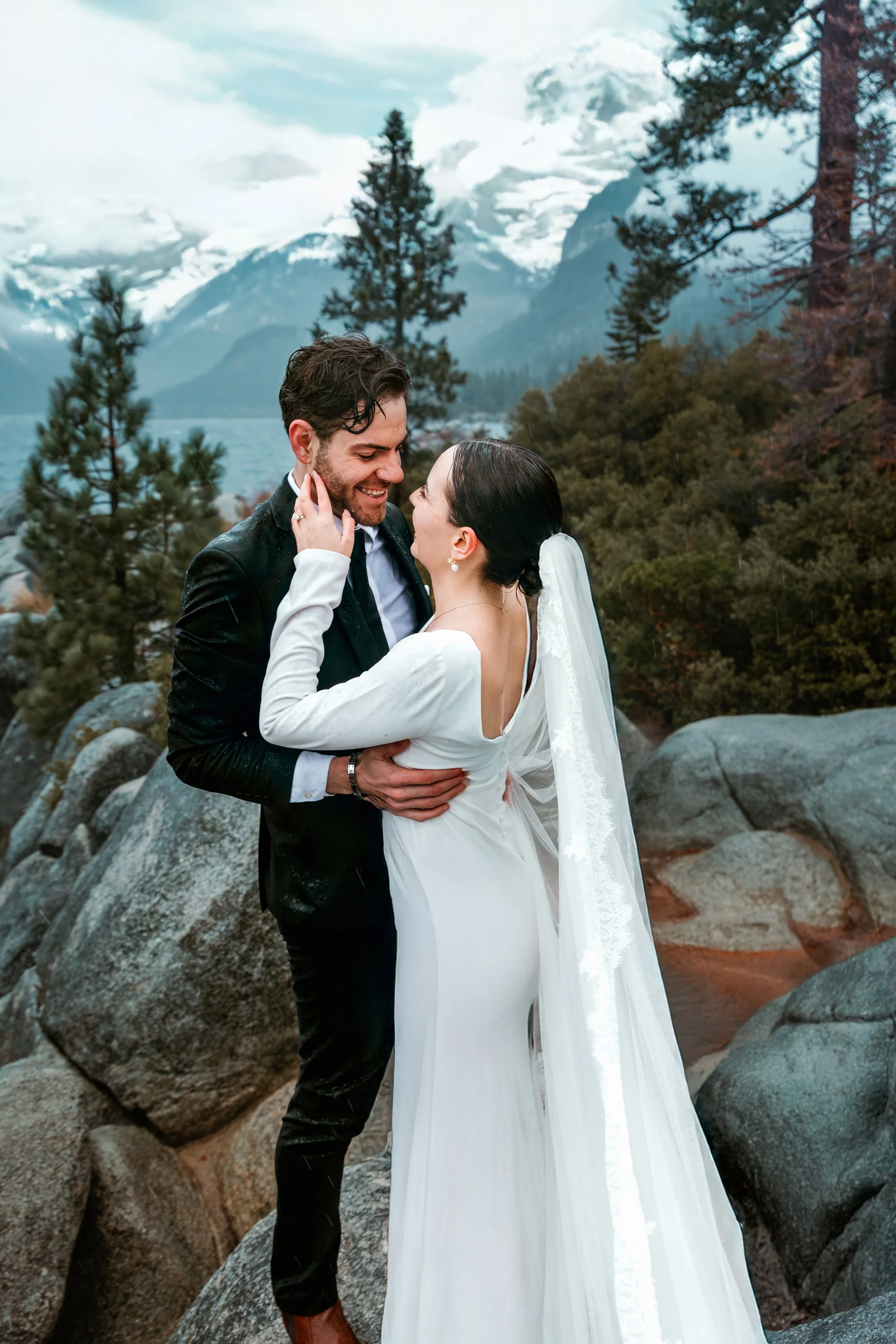 A newlywed couple in wedding attire sharing an intimate moment outdoors amidst large rocks, pine trees, and a mountainous landscape with snow-capped peaks in the background.