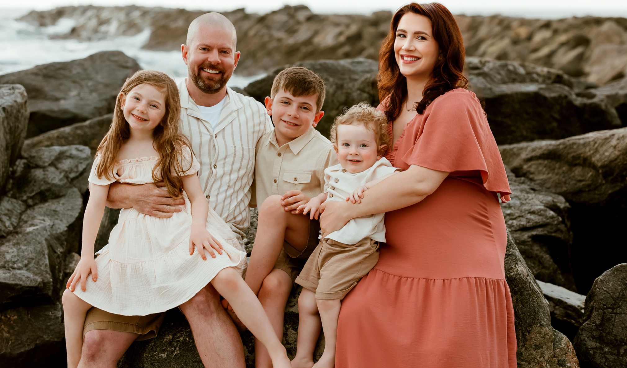 A family of six posing on rocks near the ocean during sunset, smiling at the camera. The family includes two adults and four children, with the background featuring rocks and water.