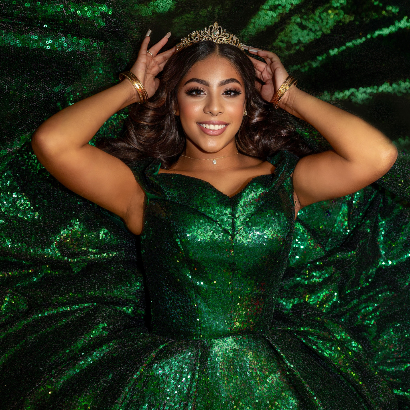 A woman wearing a green, sequined gown and a tiara, lying on a matching green, sequined fabric, smiling and holding her tiara with her hands.