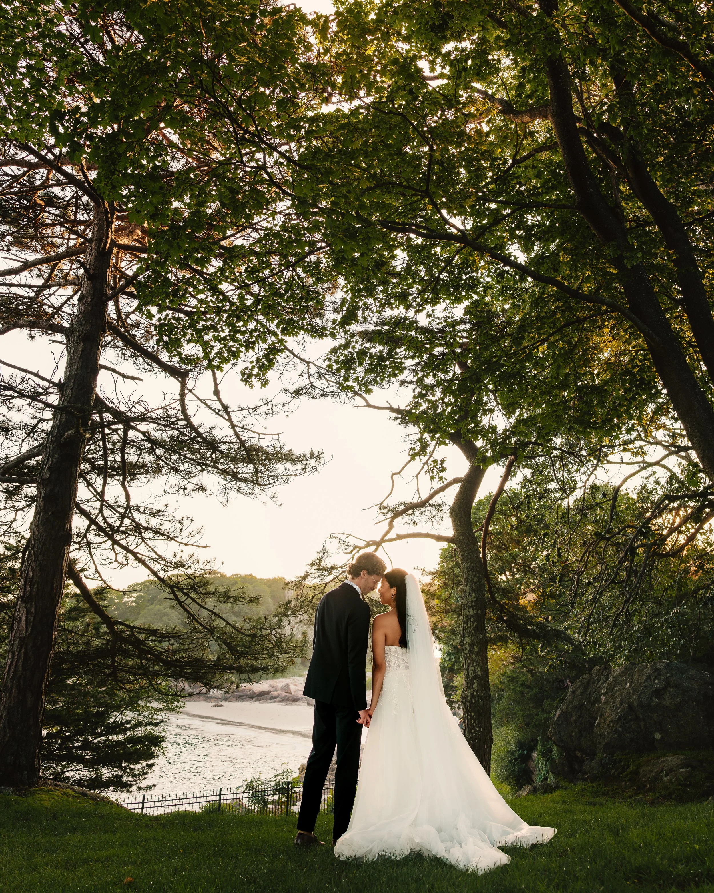 A newlywed couple standing close under trees by a river at sunset, holding hands and touching foreheads, with the bride in a white wedding gown and veil, and the groom in a black suit.
